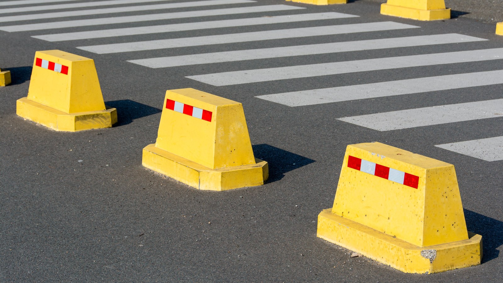 Yellow concrete traffic bollards with red-and-white reflective strips beside white zebra-crossing stripes on dark asphalt in bright daylight.