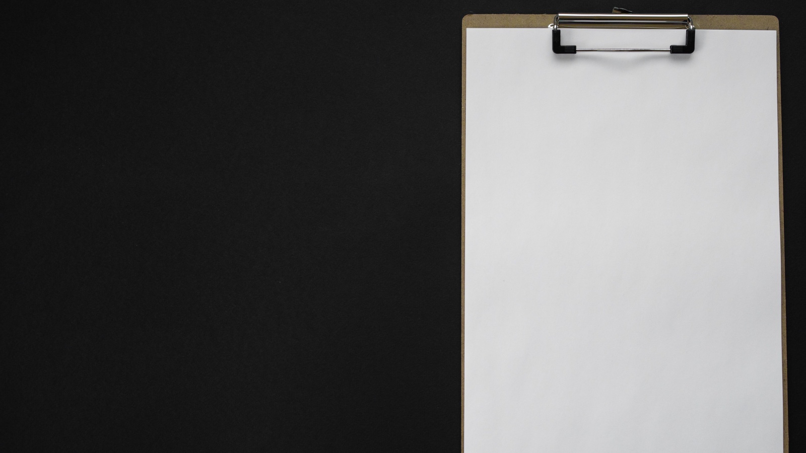 A blank white sheet of paper held by a metal clip on a brown clipboard, lying on a plain black surface.