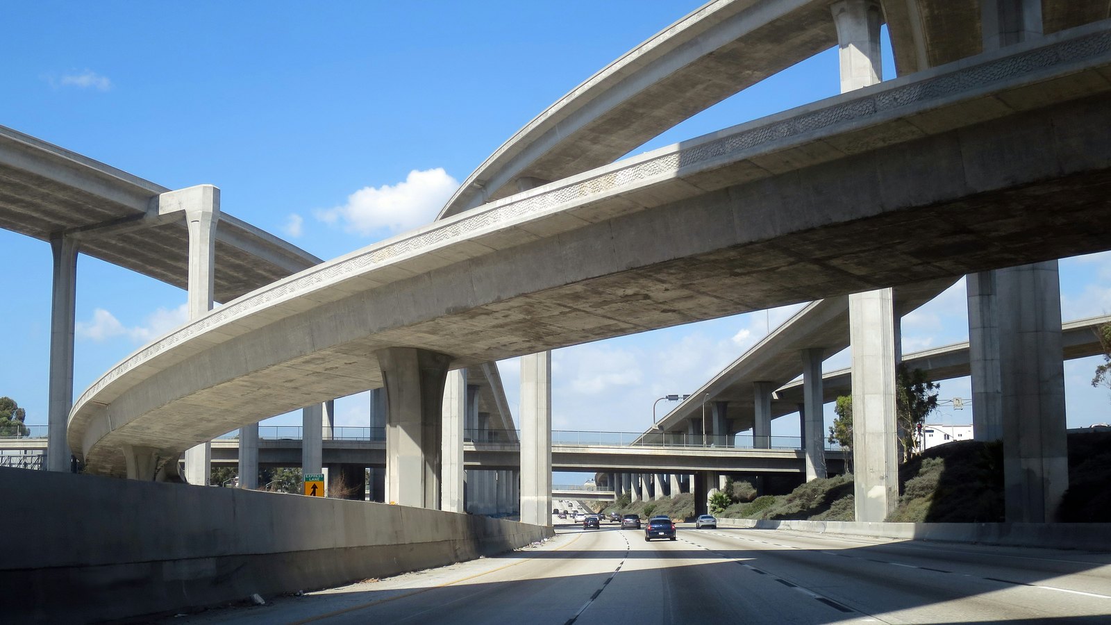 Stacked concrete freeway overpasses curving above a multi-lane freeway with a few cars ahead, under a blue sky with scattered clouds.