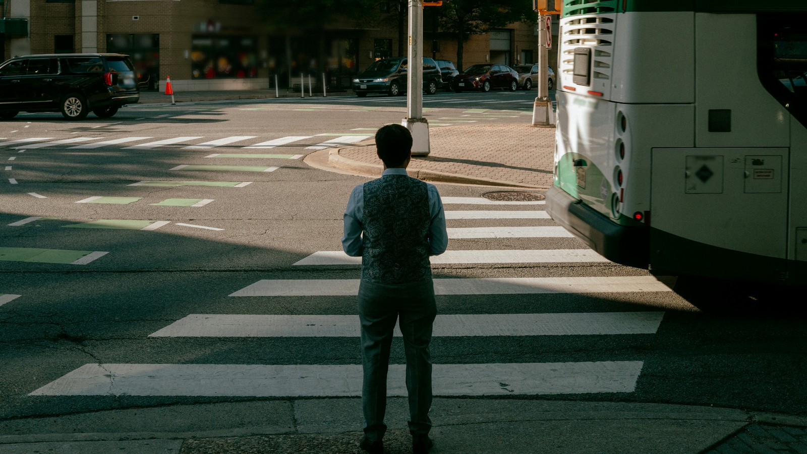 A man seen from behind stands at the curb before a striped crosswalk, a white and green transit bus beside him on the right, with a signal pole, an orange traffic cone, a black SUV, and parked cars across the intersection in front of a brick building with storefronts.