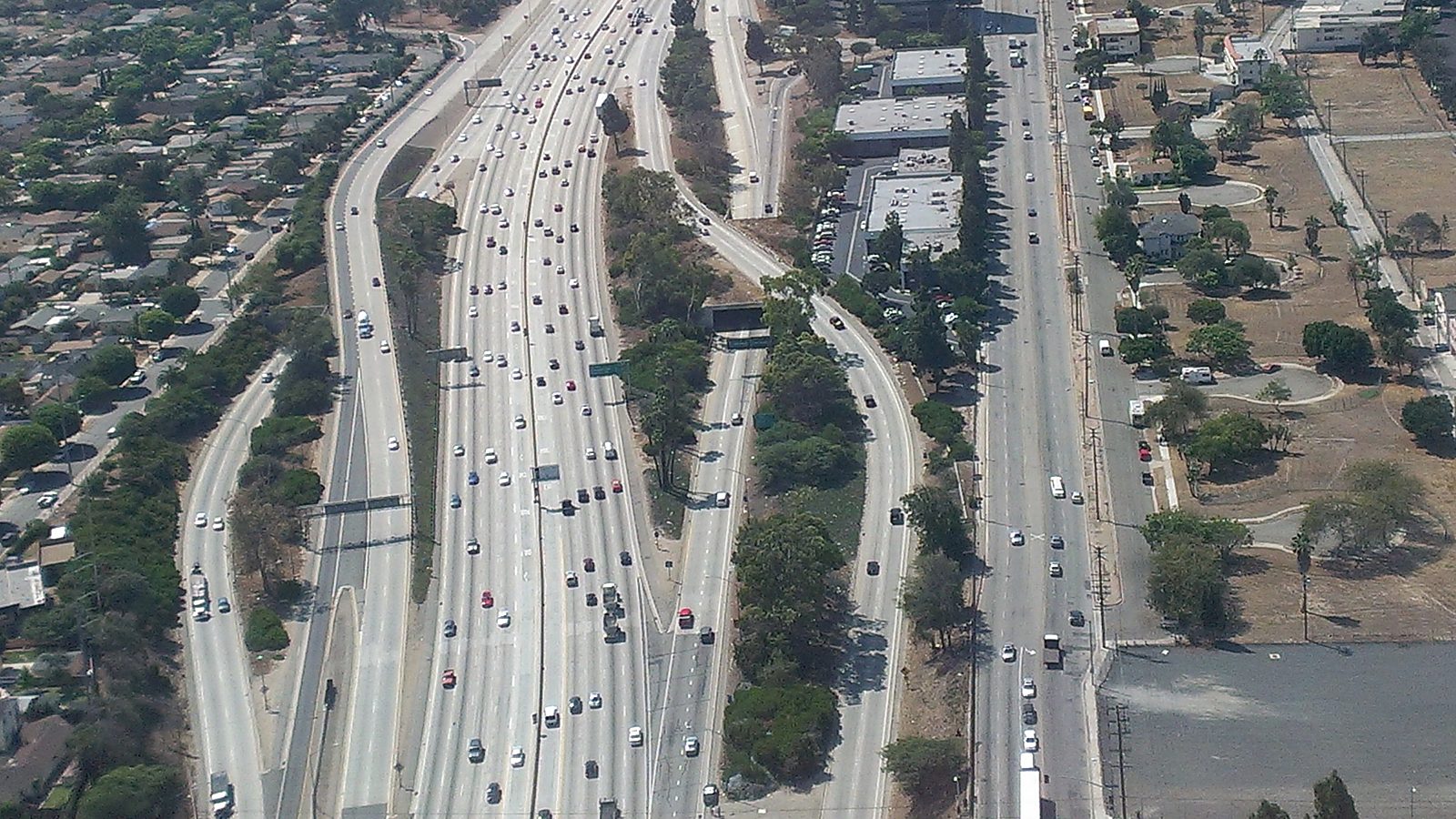 Aerial view of a wide Los Angeles-area freeway packed with small cars heading in both directions, with on-ramps, a frontage street, rows of houses, trees, and low commercial buildings alongside.