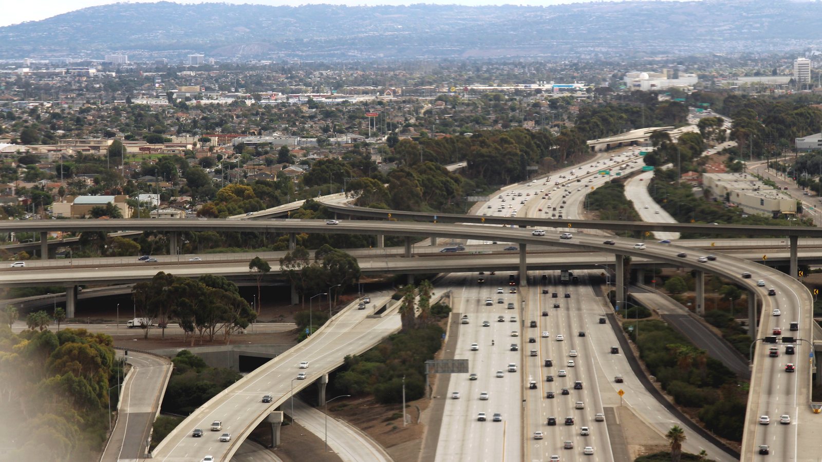 Aerial view of a large freeway interchange with stacked curving overpasses and a multi-lane freeway full of small cars, with neighborhoods, trees, and hills in the distance.
