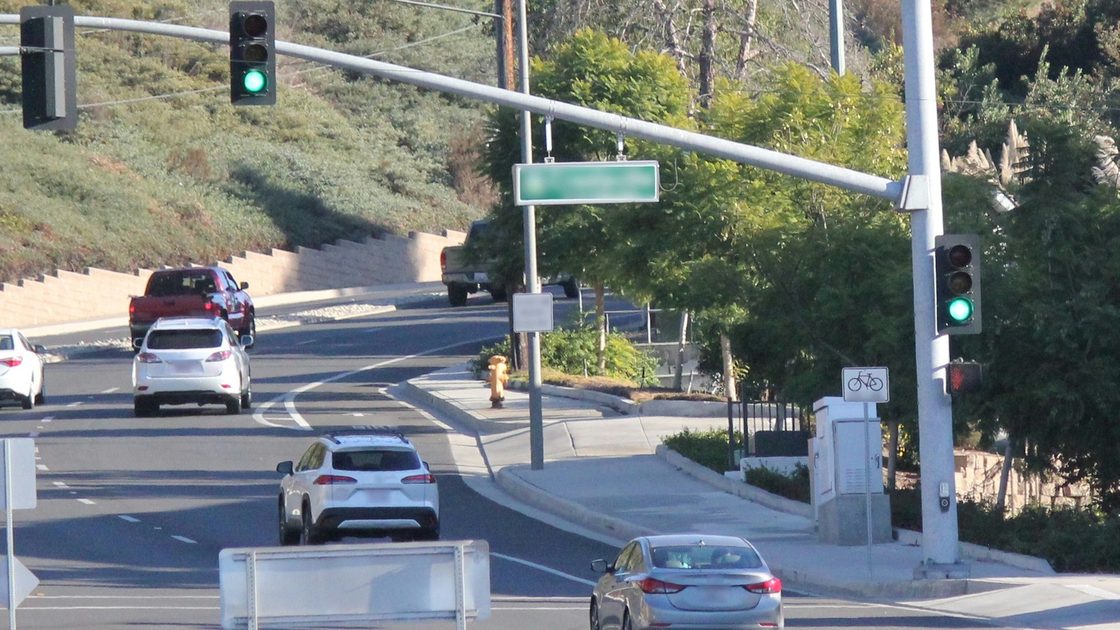Cars travel a curving multi-lane road at a signalized intersection, with green traffic lights on a mast arm and a corner signal pole, a red pickup and two white SUVs beside a hillside retaining wall, and a silver sedan in the near lane.