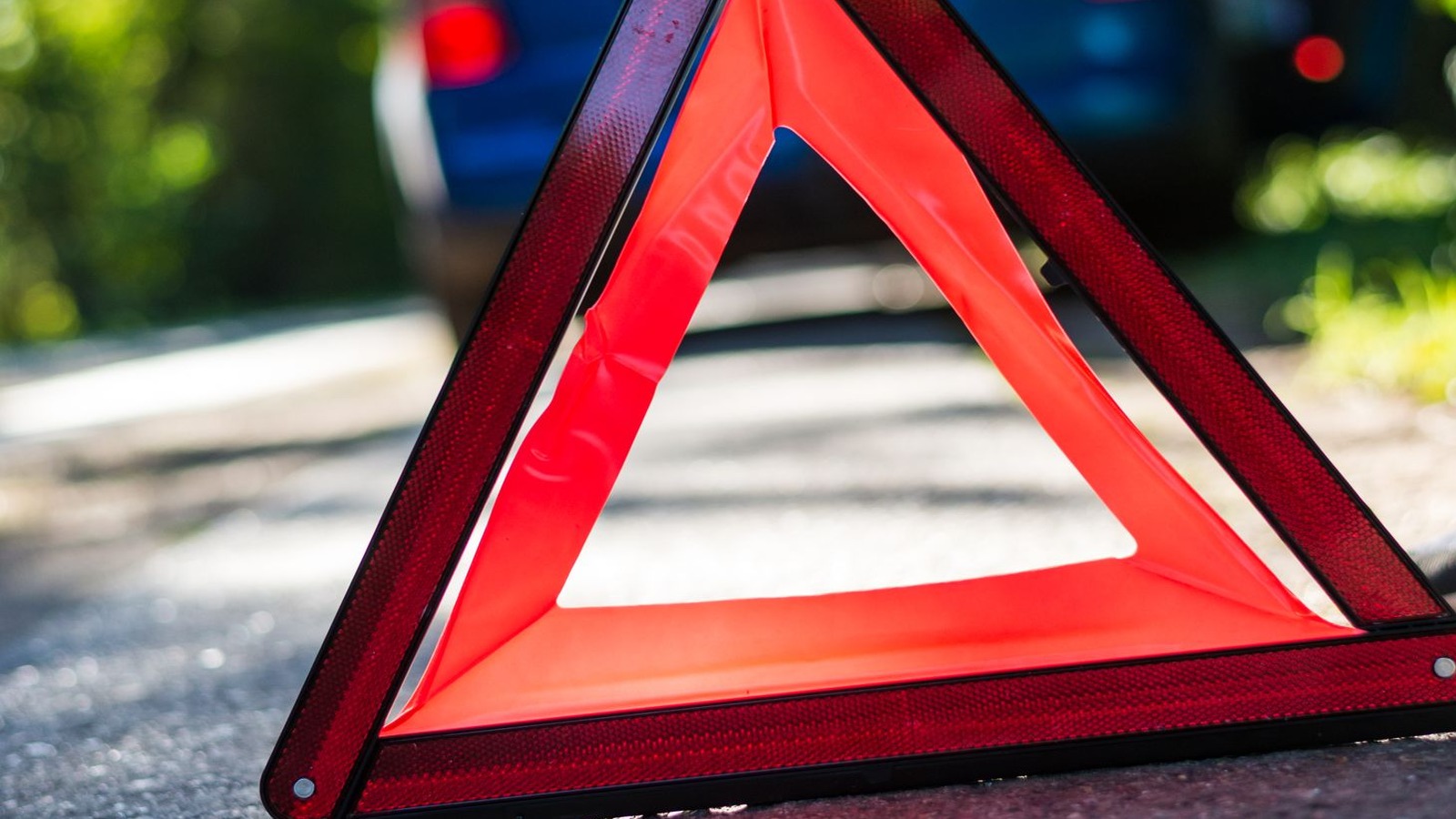 A red reflective emergency warning triangle on asphalt in front of a blurred blue car, with soft-focus green roadside foliage in daylight.