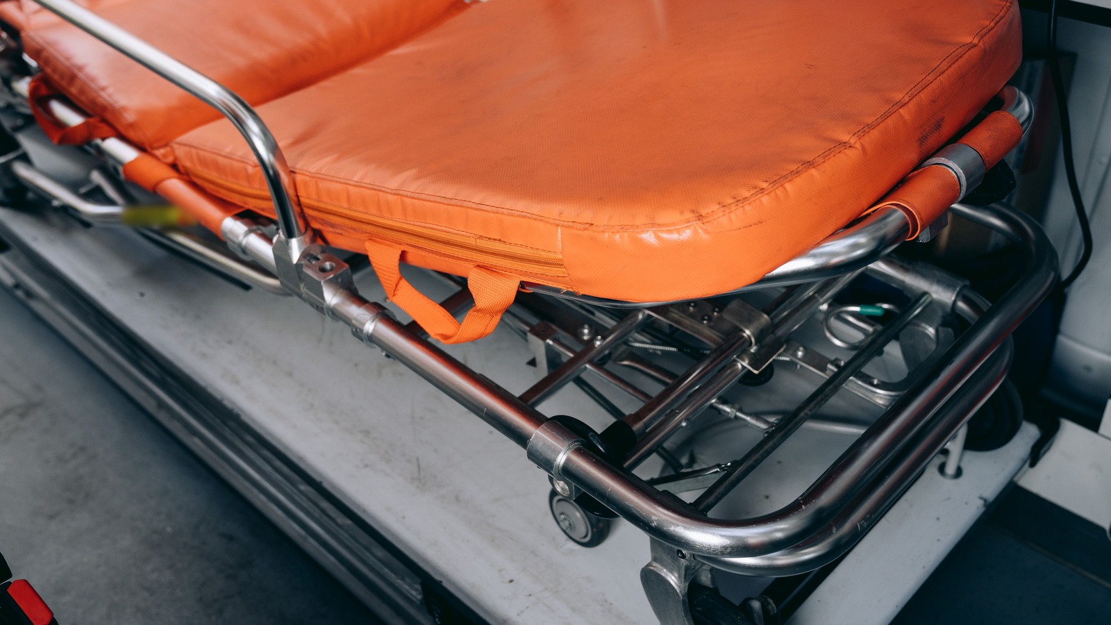 An empty orange padded stretcher with a chrome frame, carry straps, and small wheels, resting on a light gray floor.