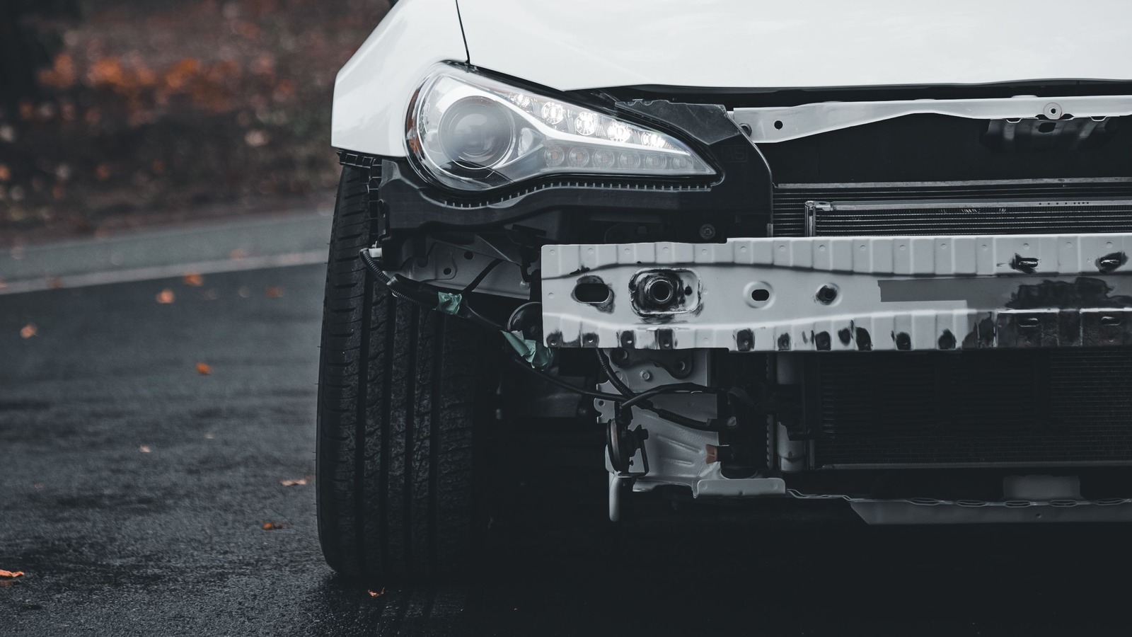 Close-up of the front of a white car with the bumper cover torn away, exposing a bare metal reinforcement bar, loose wiring, and the radiator, next to an intact headlight and front tire on dark asphalt with a few fallen leaves.