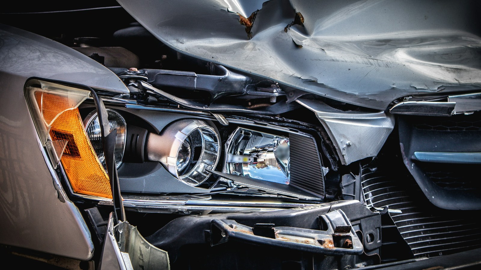 Close-up of a silver car's damaged front end, with a crumpled and creased hood showing rust at the folds, a cracked amber corner lamp, an exposed headlight assembly, and a bent bumper bracket.