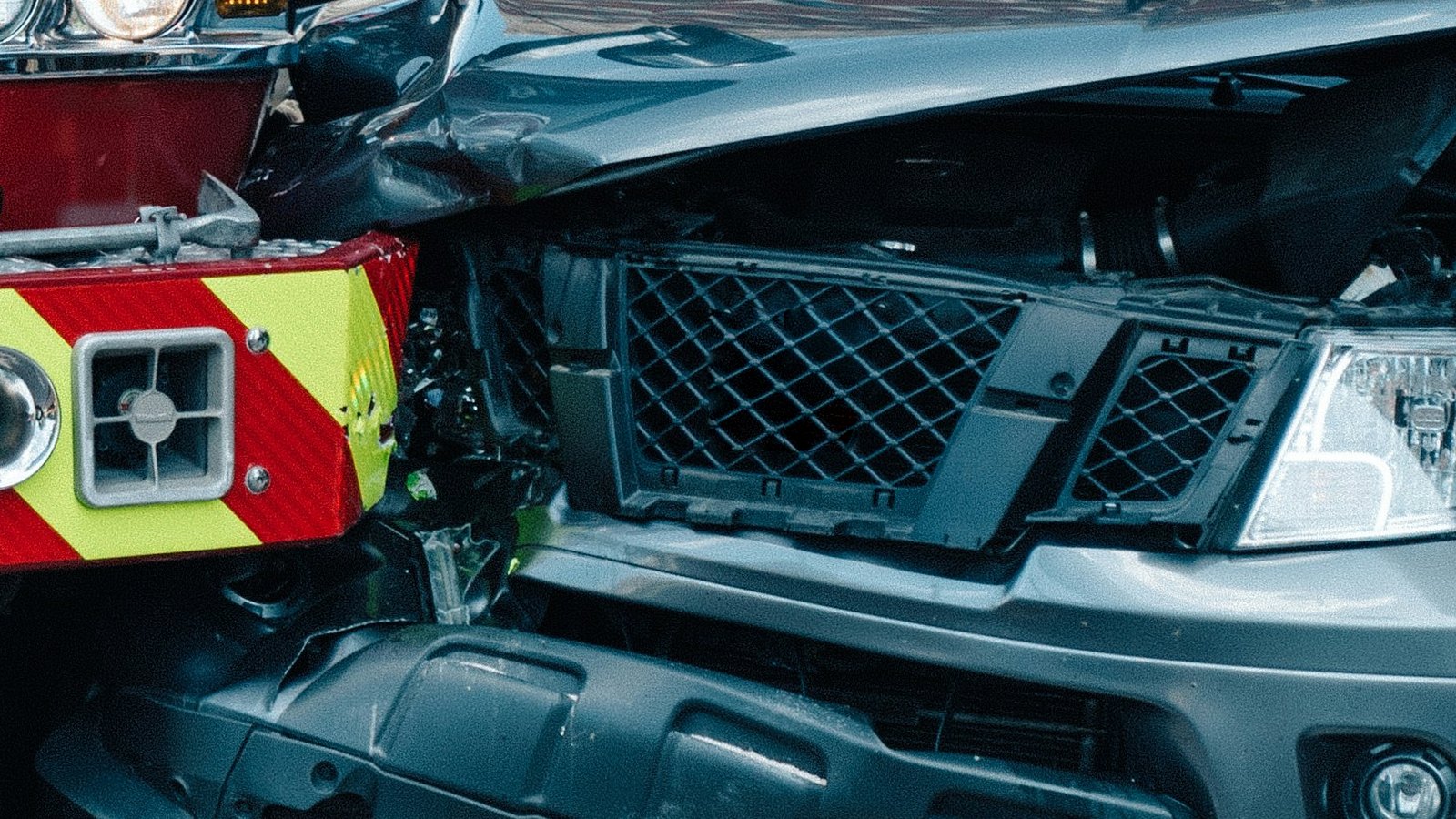 Close-up of the crumpled hood and bent grille of a gray pickup truck pressed against the red and yellow striped front bumper of a large red truck, with a cracked headlight and a twisted lower bumper on the pickup.