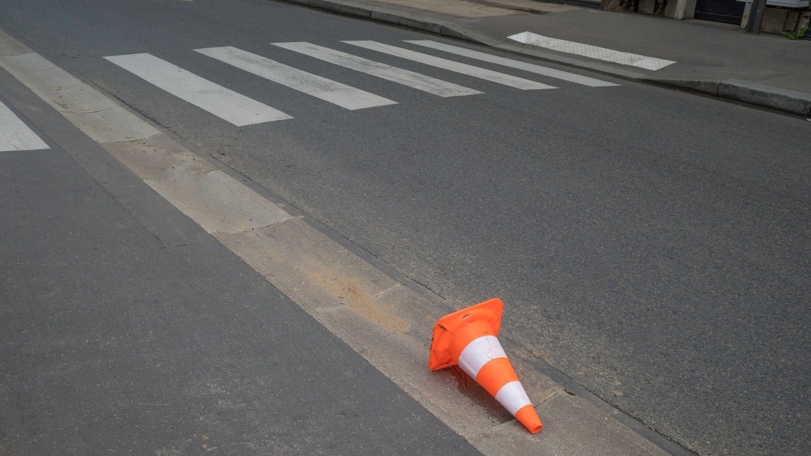 An orange traffic cone with white reflective bands lying on its side in a concrete gutter beside a zebra crosswalk on a gray asphalt street.