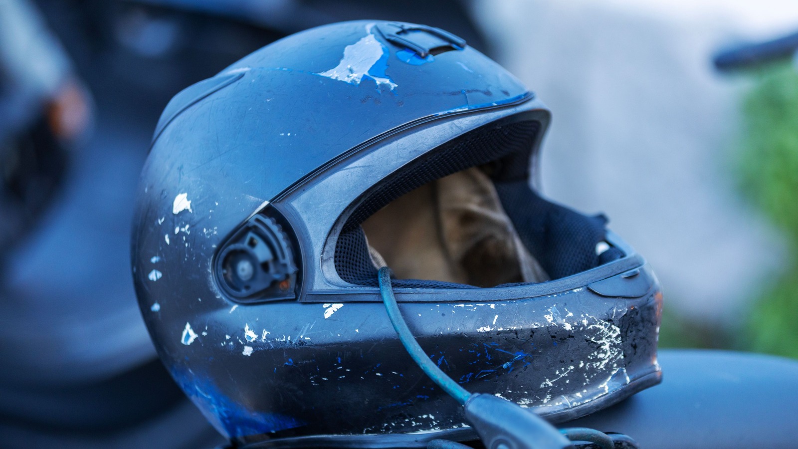 A scuffed dark blue full-face motorcycle helmet with chipped paint and no visor, resting on a motorcycle seat with a cable lock looped through it, against a softly blurred background.