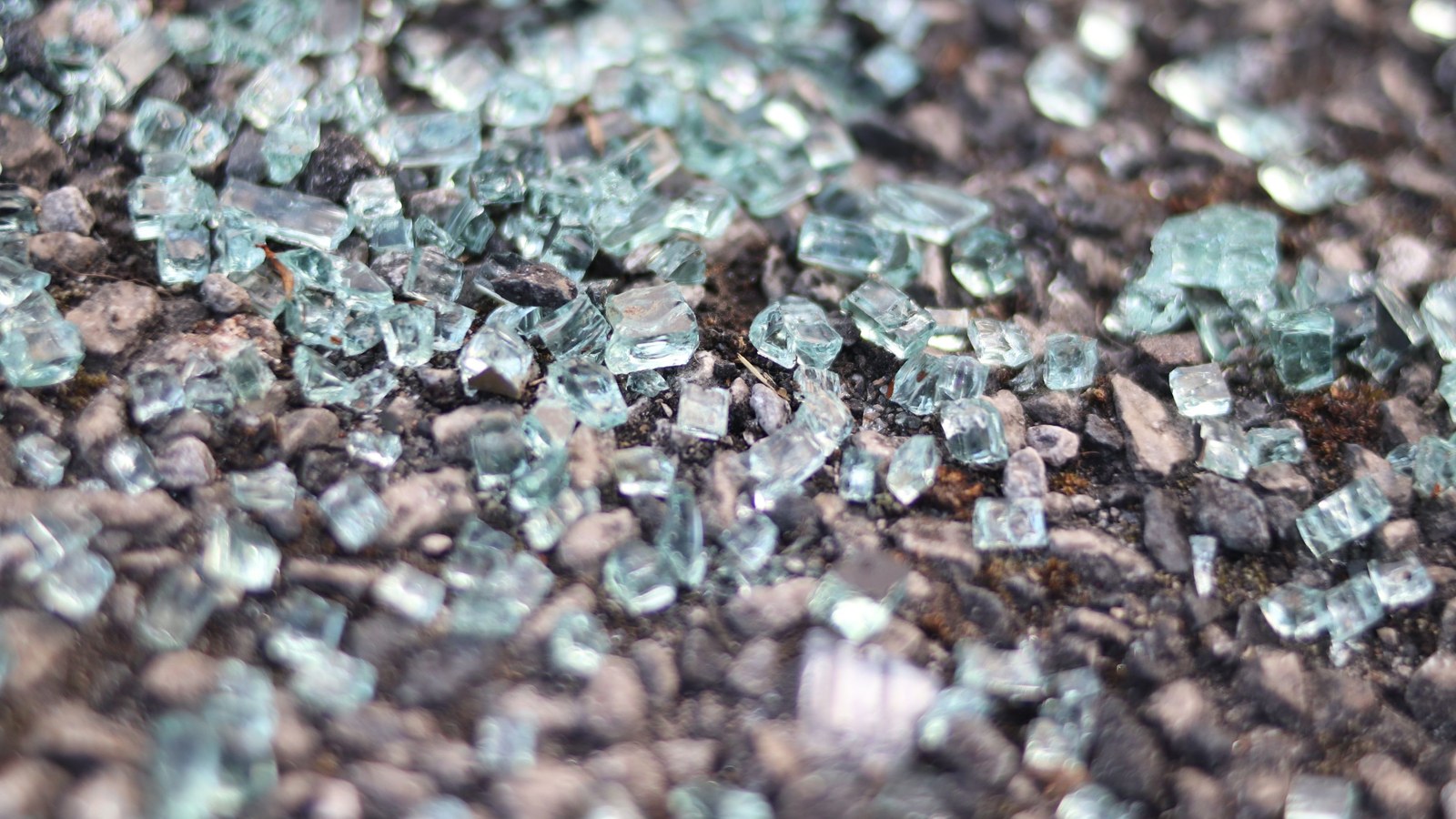 Macro close-up of pale blue tempered-glass cubes scattered on dark textured asphalt and gravel in soft daylight.