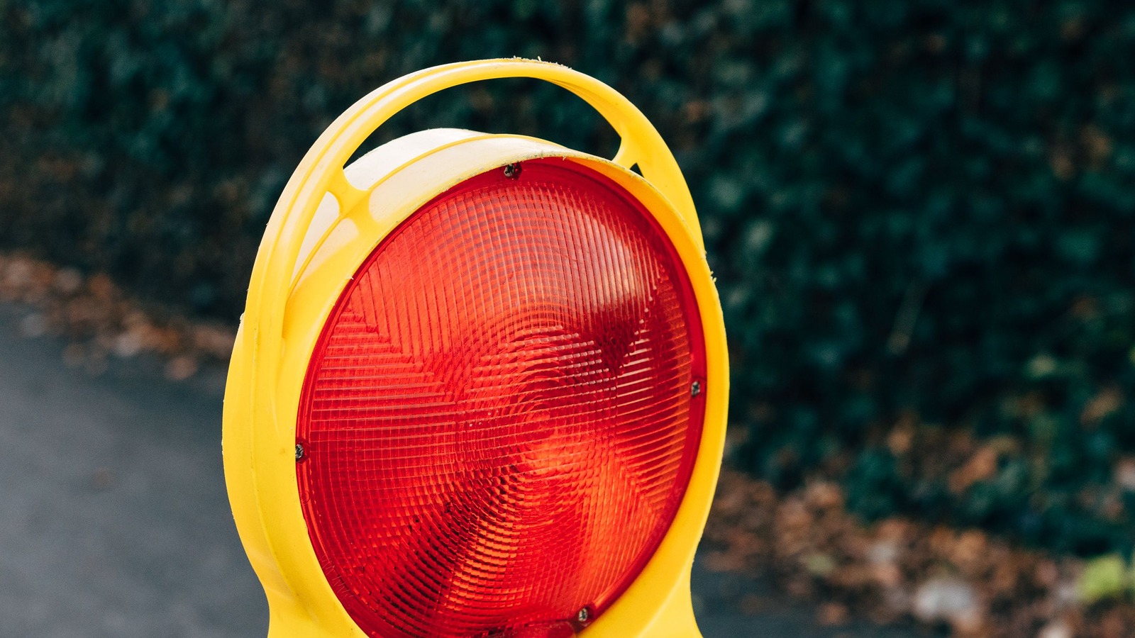 Close-up of a red circular warning light in a yellow housing on a road construction barrier beside asphalt and dry leaves.