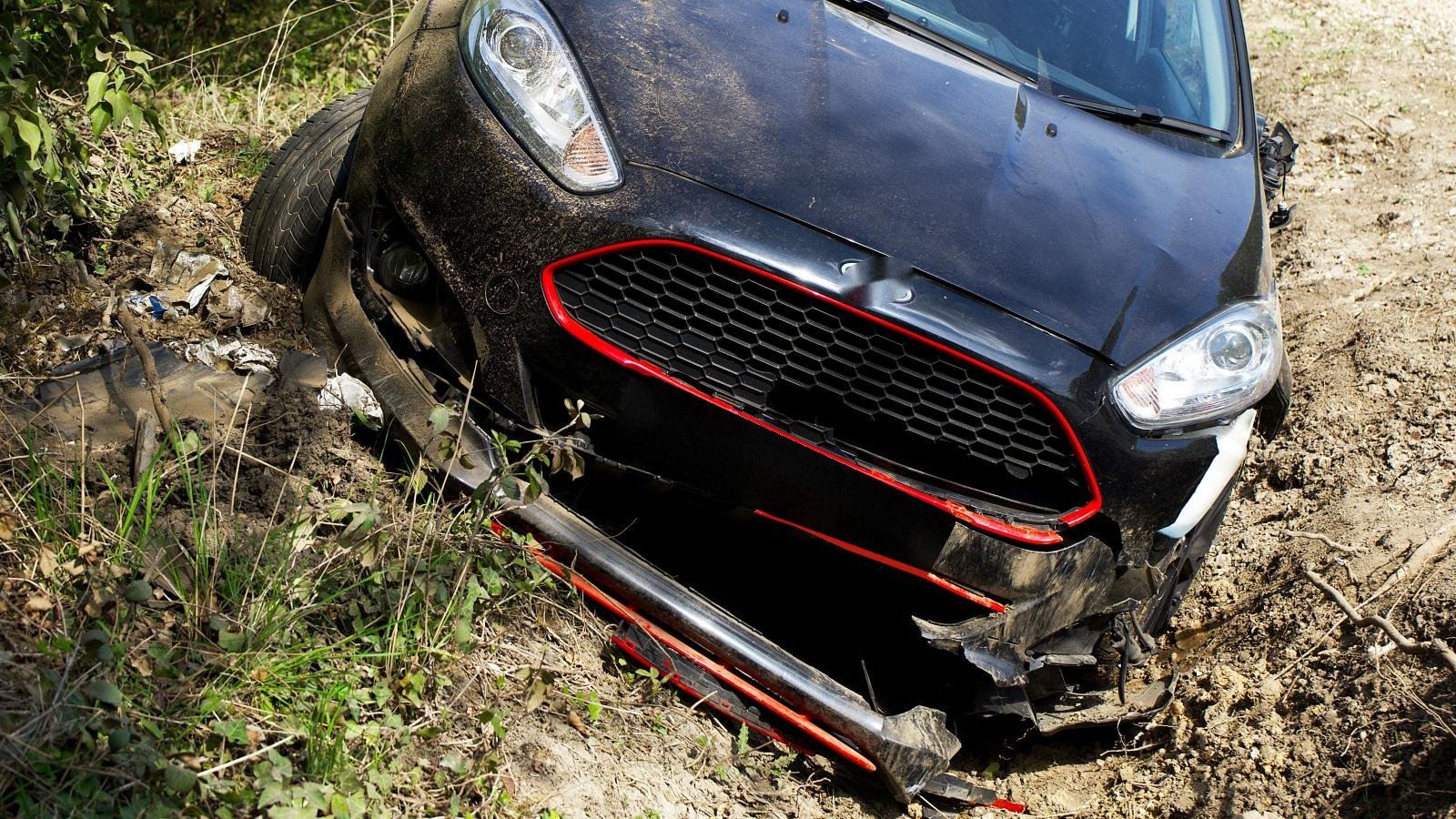 A black hatchback nosed down into a dirt roadside ditch, its front bumper torn loose and hanging to the ground, with broken trim and plastic pieces scattered under the front corner.