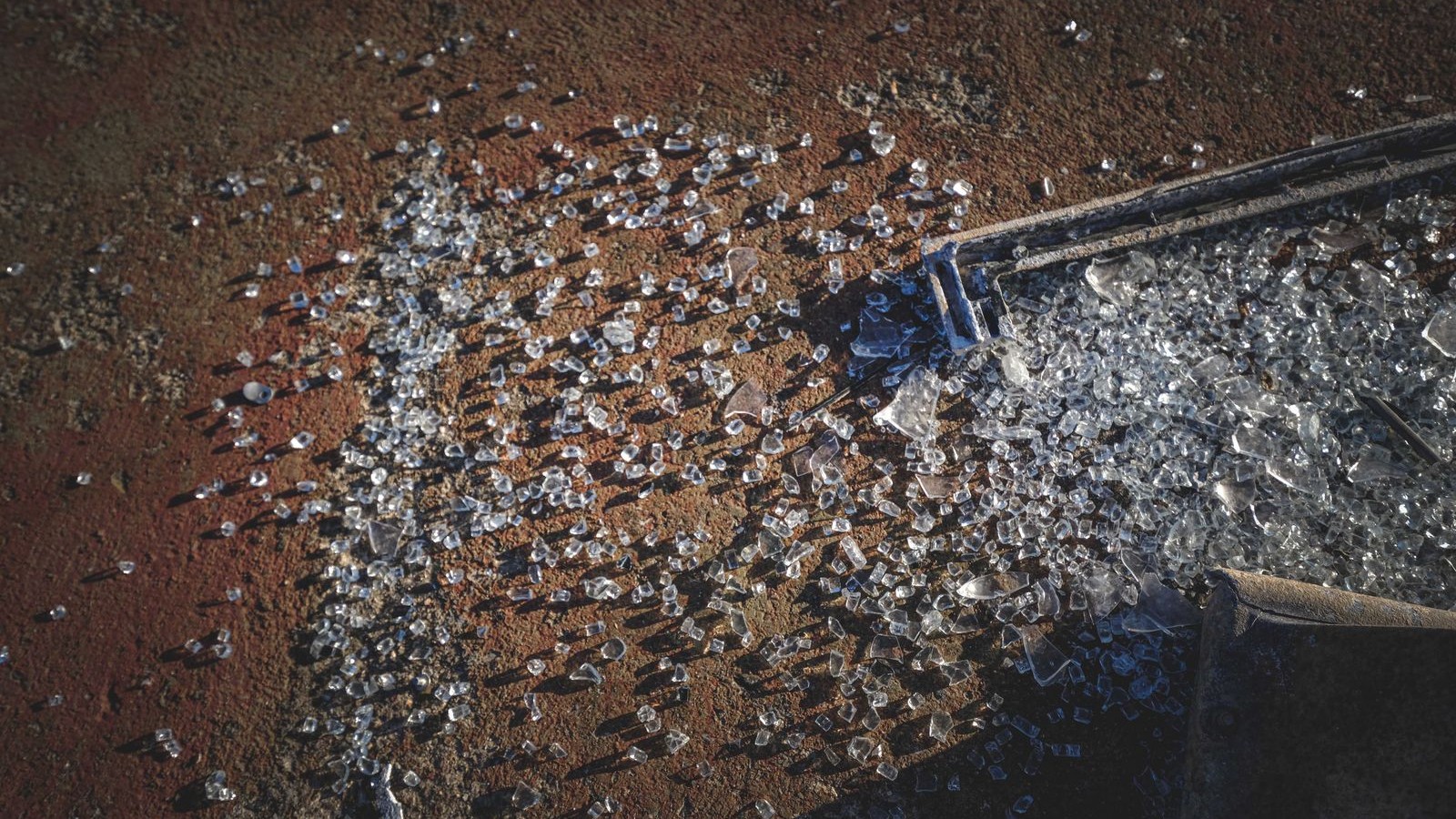 Close-up of clear shattered tempered-glass shards and dark trim debris scattered on rough reddish-brown asphalt in bright daylight.