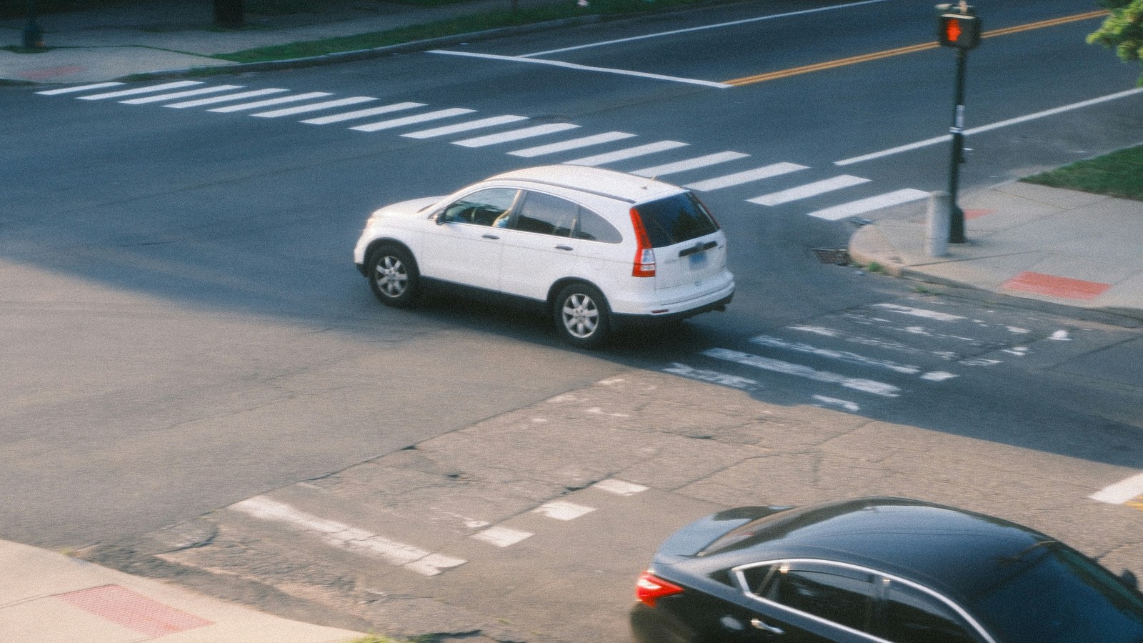 A white SUV turning through a street intersection beside a white-striped crosswalk, with a double yellow center line beyond it, a pedestrian signal showing a red hand on the corner, and the roof of a dark sedan in the foreground.