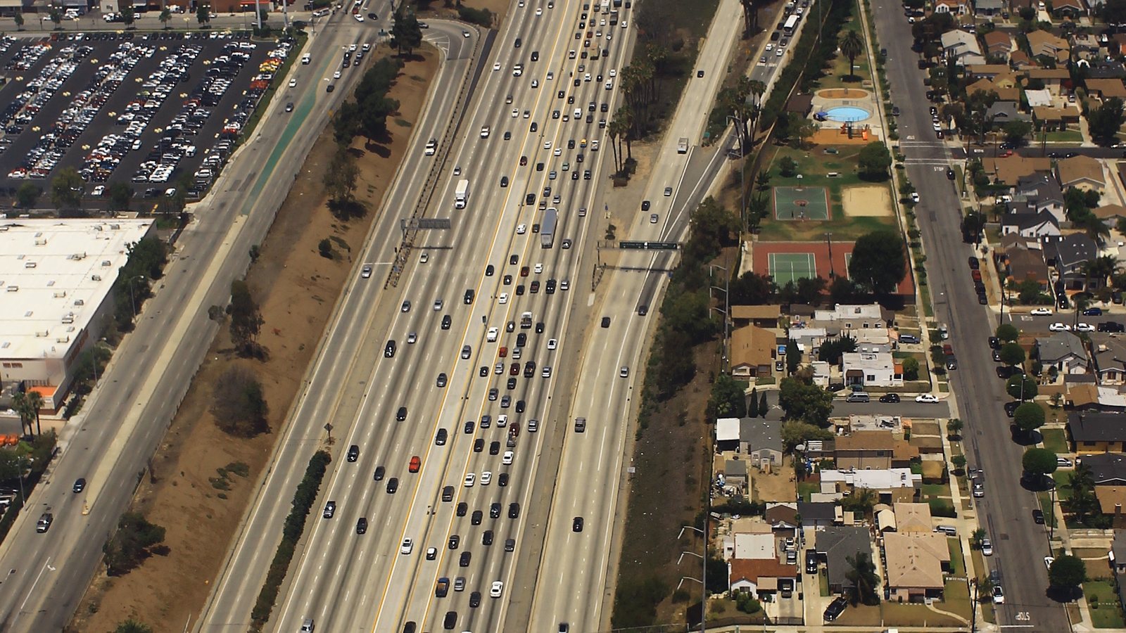 Aerial view of a wide Southern California freeway packed with cars in both directions, with frontage roads, a large flat-roofed building and parking lot on one side, and neighborhoods of houses, trees, and a small park on the other.
