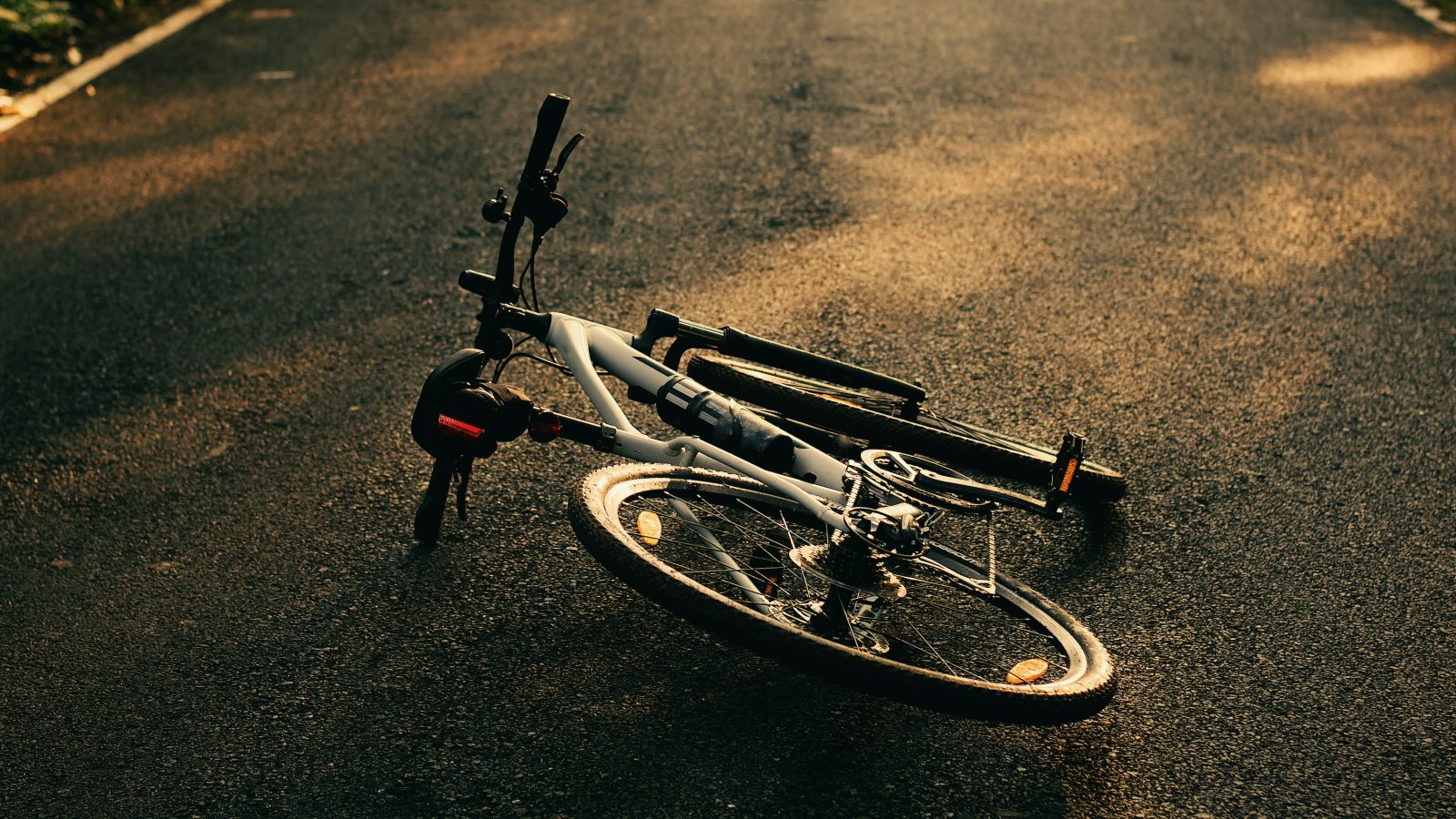 A white mountain bike lying on its side in the middle of an empty asphalt road in warm, low sunlight, with a white edge line and a strip of greenery in the top corners.