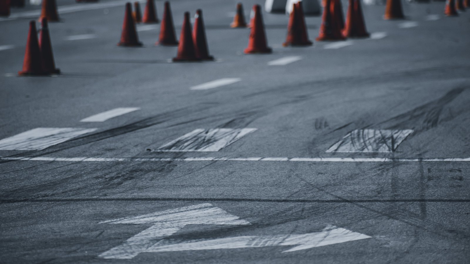 Dark tire skid marks curve across gray asphalt over white painted arrows and lane lines, with rows of orange traffic cones blurred in the background and no people, text, or logos visible.