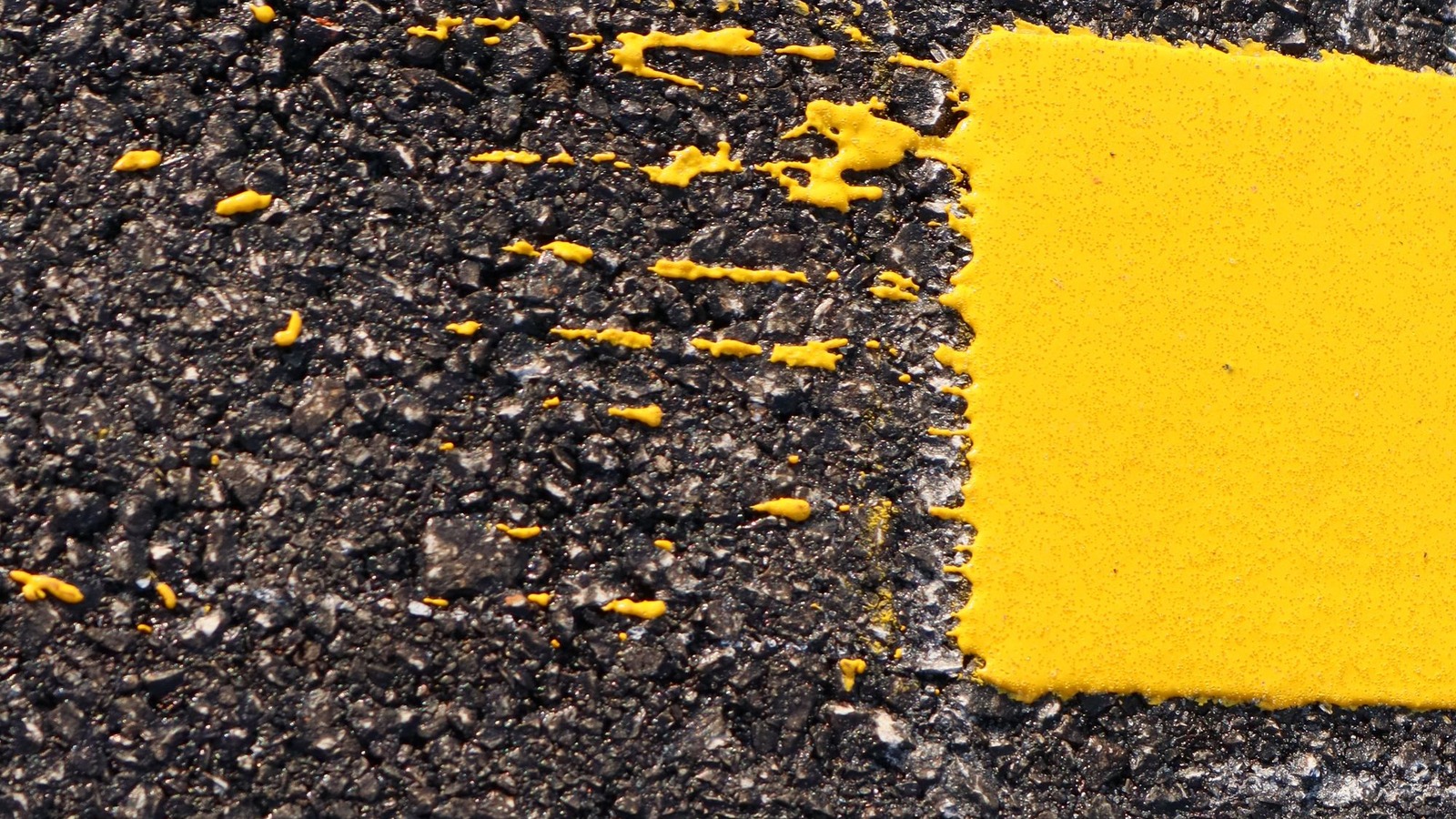 Close-up of fresh yellow road paint with irregular overspray streaks on coarse dark asphalt.