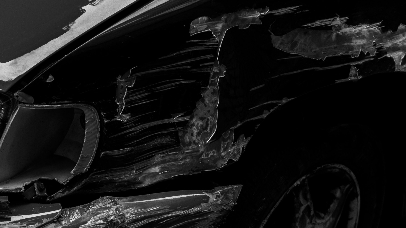 Black and white close-up of a dark car's front fender and bumper with long scrapes and peeled paint, a broken turn signal housing, and the top of a front wheel.