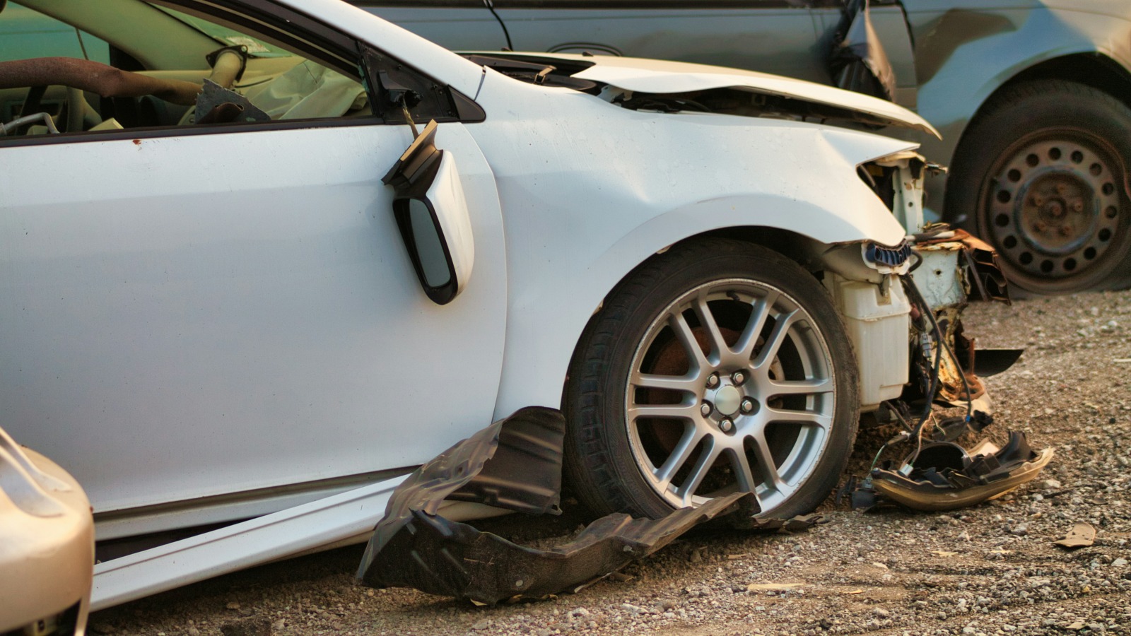 The crushed front corner of a white car parked on gravel, with its bumper torn away, the fender bent and a black plastic wheel liner lying on the ground beside the front wheel, next to another damaged car.