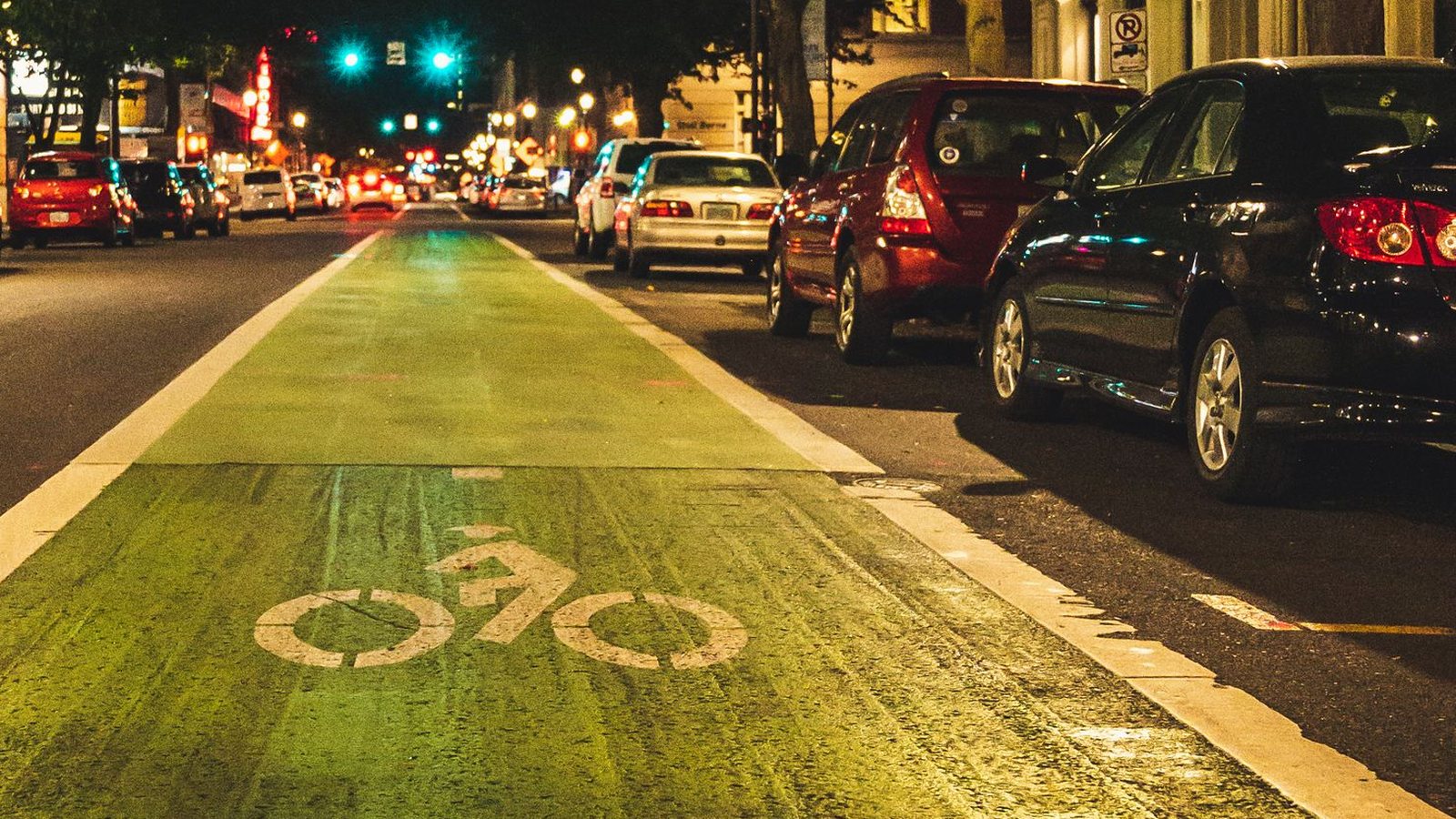 A green-painted bicycle lane with a white bicycle symbol runs down a city street at night, between a traffic lane and a row of parked cars, with green traffic signals in the distance and no people visible.