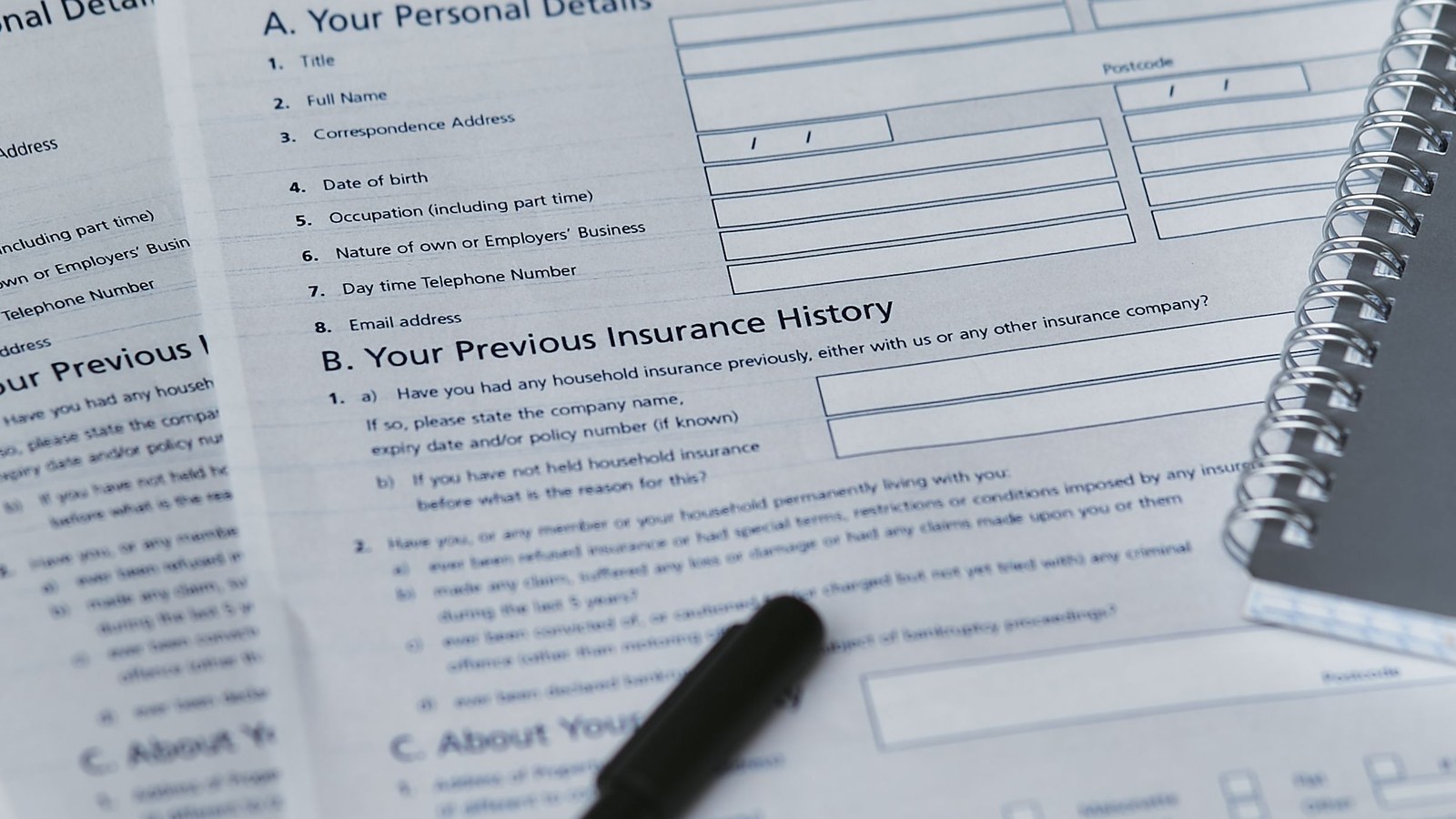 Blank insurance claim forms for personal details and previous insurance history on a desk, with a black pen and a spiral notebook.