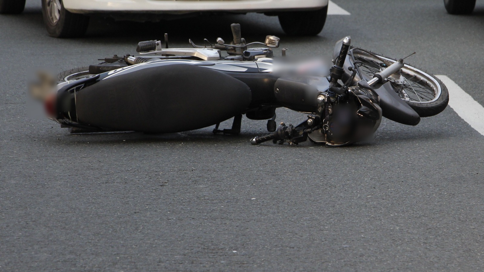 Una motocicleta negra tirada de lado sobre el asfalto gris, con un casco junto al manubrio, la rueda delantera cerca de una línea blanca del carril y, al fondo, la parte baja de un auto blanco detenido.