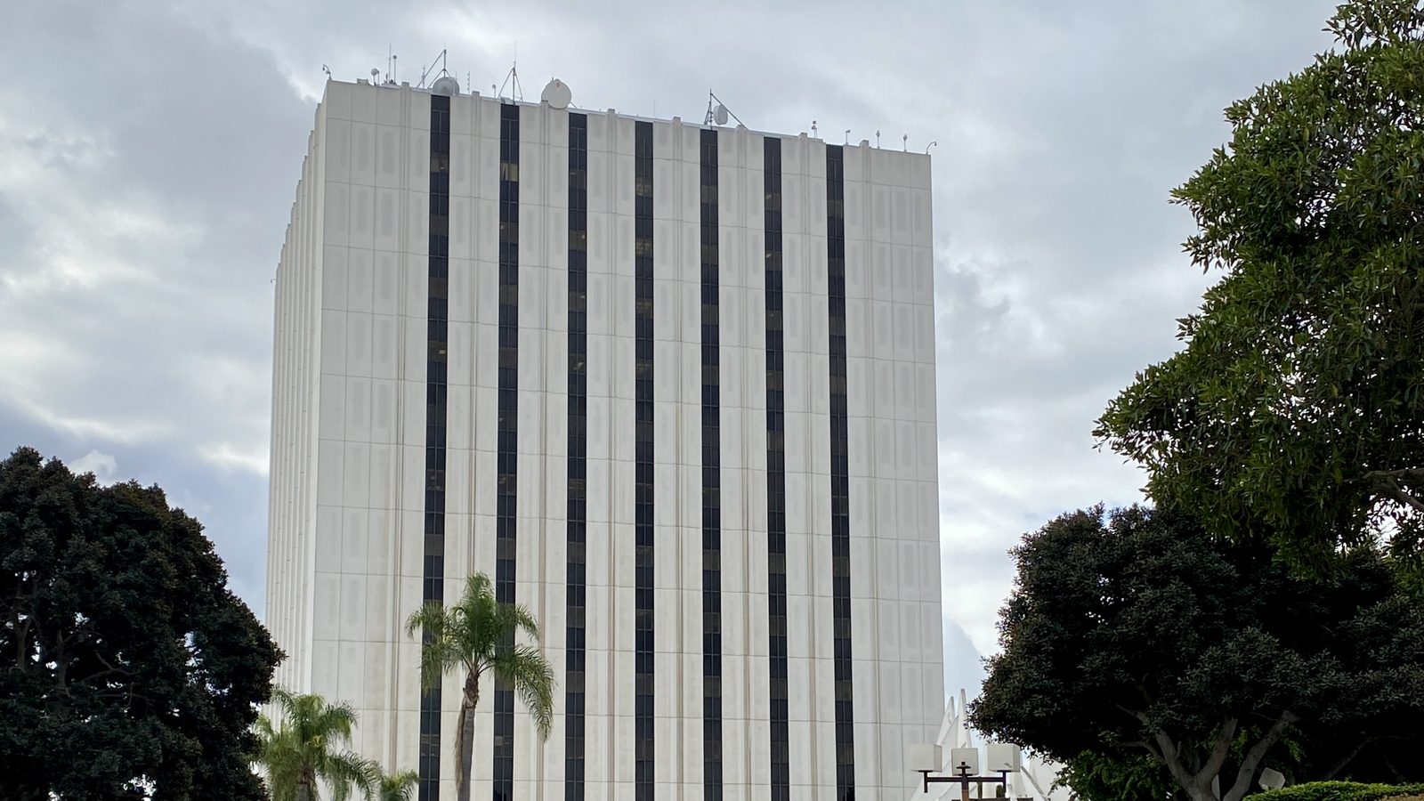 Torre blanca de un juzgado con franjas verticales de ventanas oscuras y antenas en la azotea, con palmeras y árboles al frente, bajo un cielo nublado.