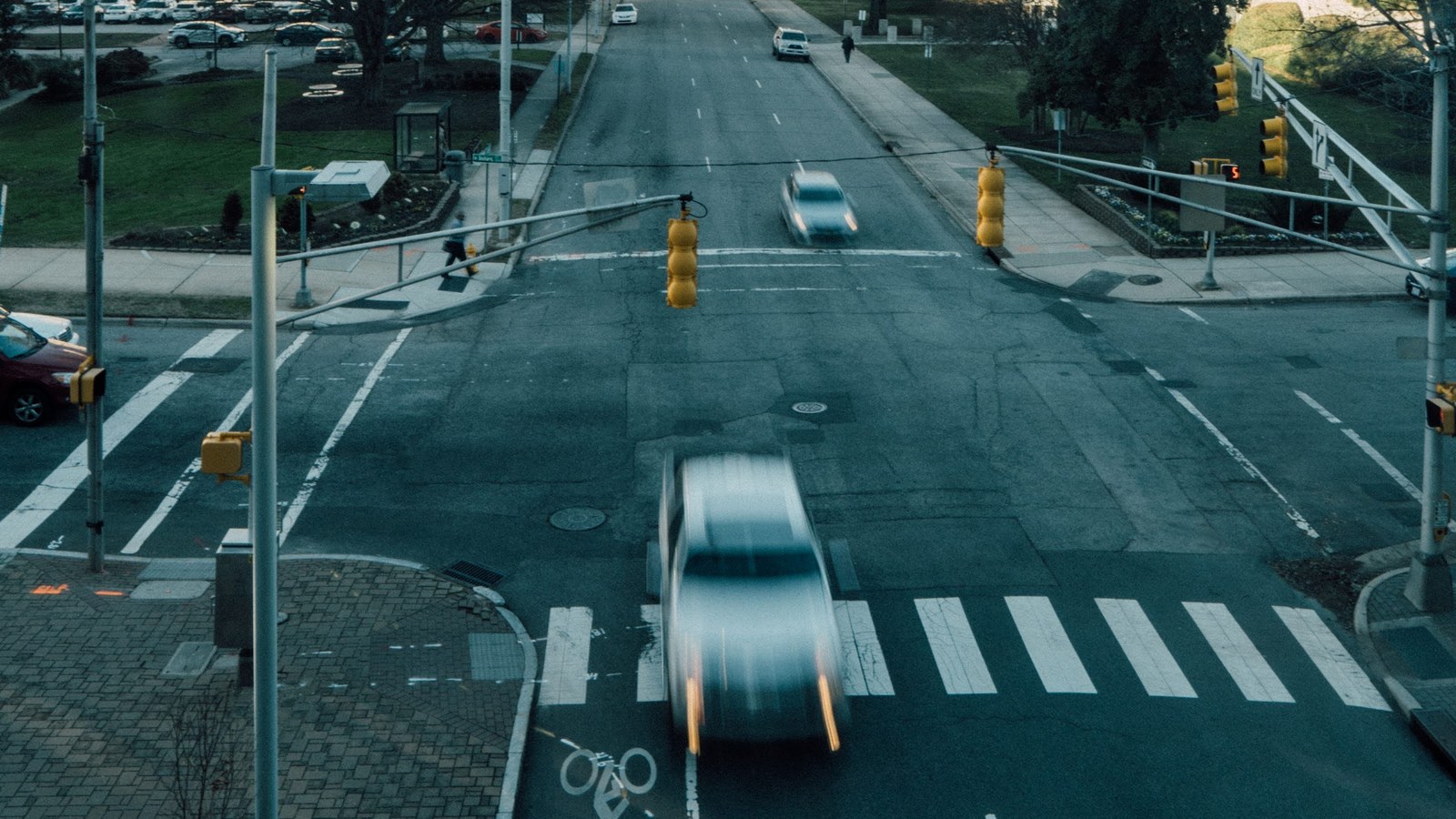 Vista elevada de un cruce con semáforos y paso peatonal, con un auto en movimiento que se ve borroso y otros autos en la calle al fondo.