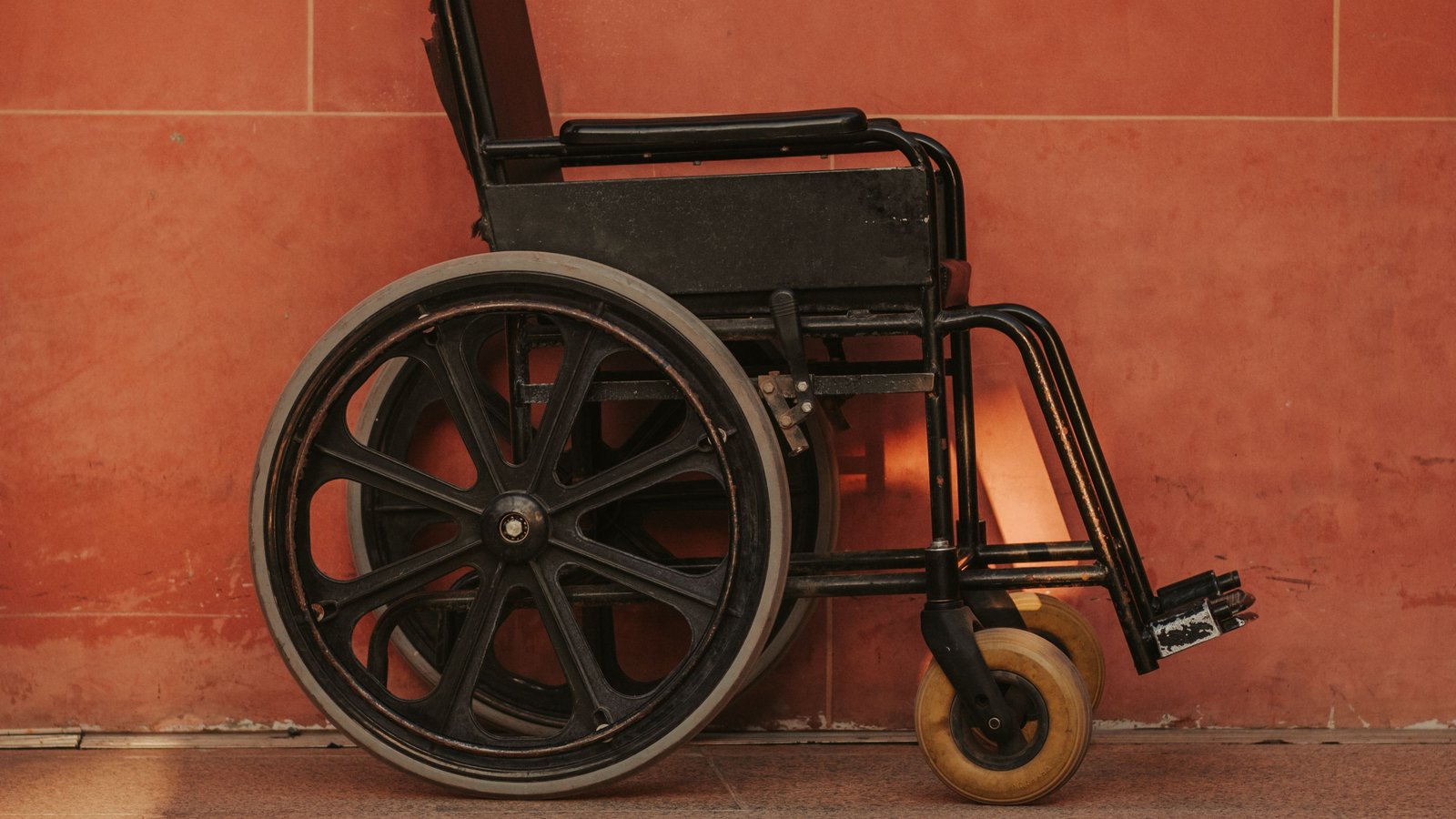 Una silla de ruedas negra y vacía, vista de lado, junto a una pared color terracota, sobre un piso de baldosas; no hay personas, texto ni logotipos.