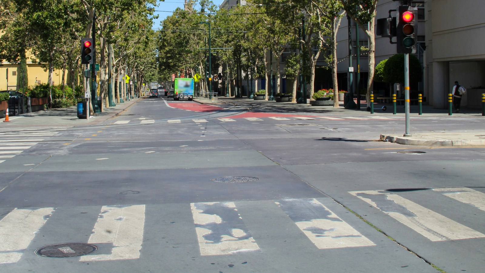 Intersección amplia con pasos de peatones blancos en primer plano, semáforos en rojo, una avenida bordeada de árboles al fondo con vehículos a lo lejos y una persona que camina por la acera de la derecha.