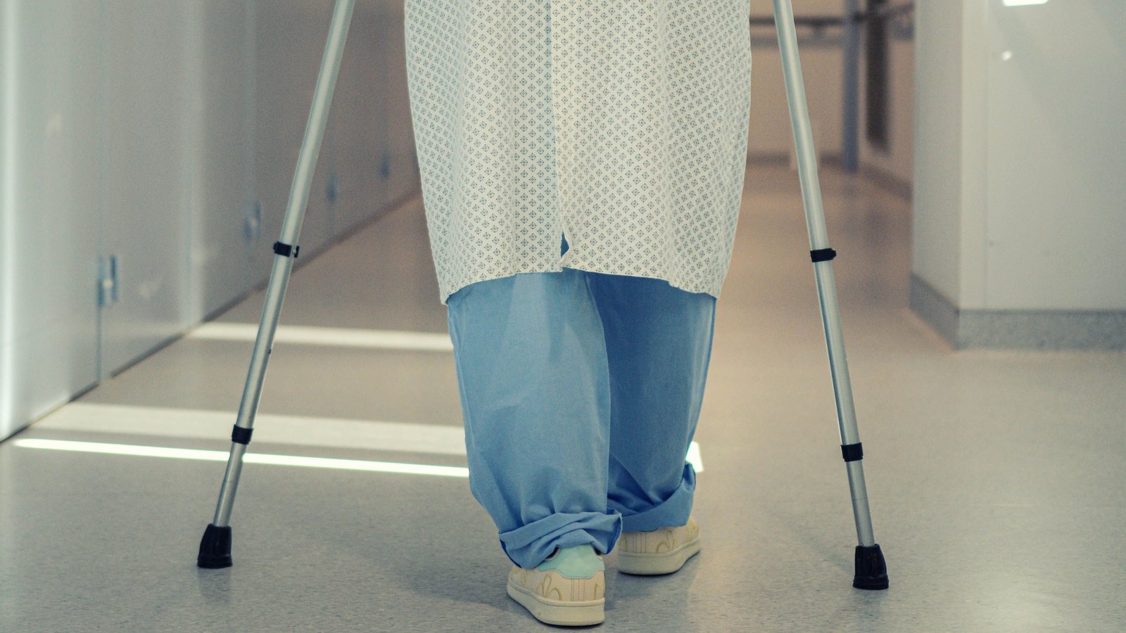 Vista de espaldas, de la cintura para abajo, de un paciente con bata de hospital que camina con dos muletas por un pasillo de hospital; no se ve la cara ni hay texto ni logotipos.