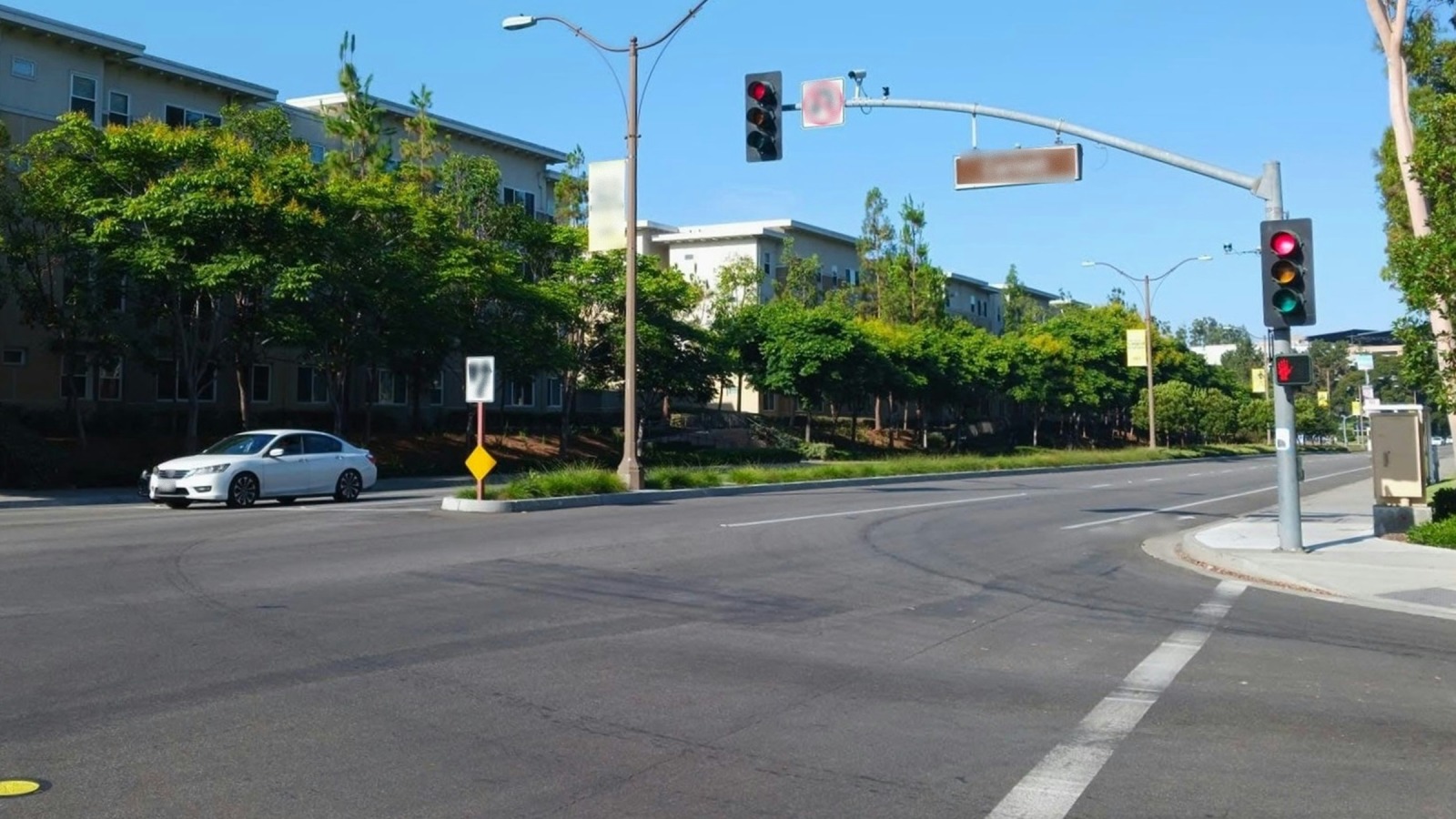 Un sedán blanco cruza una amplia intersección con semáforos en rojo, una señal peatonal con la mano roja, una franja blanca pintada en el asfalto y edificios de apartamentos de pocos pisos detrás de árboles verdes bajo un cielo despejado.