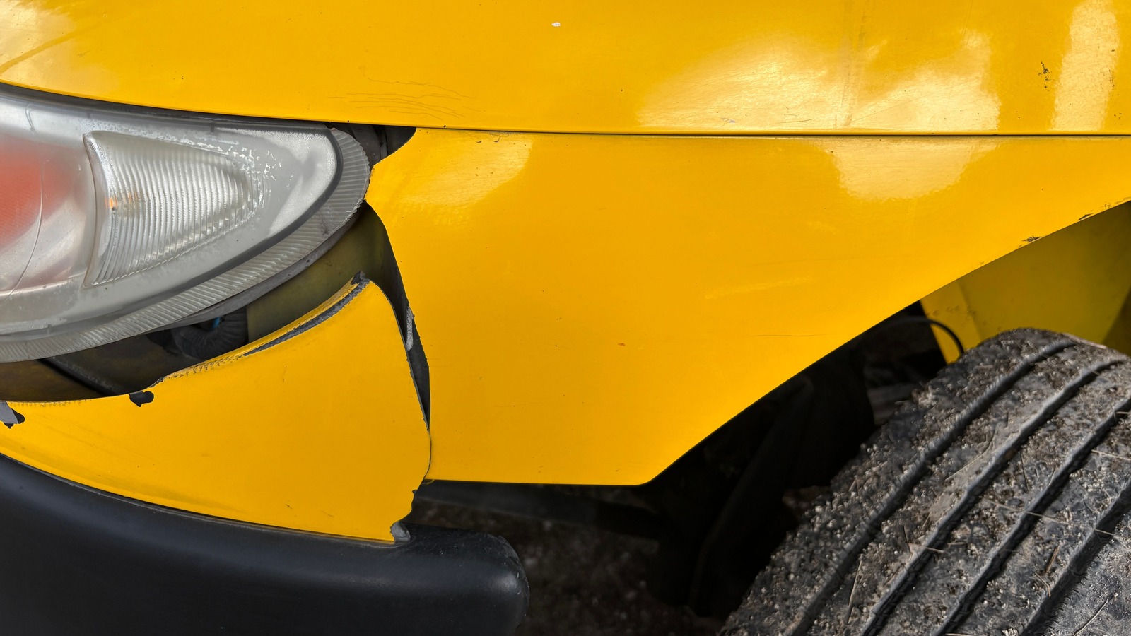 Primer plano de la esquina delantera de un auto amarillo, con la cubierta de la defensa rota y separada de la salpicadera, el borde del faro desajustado, la defensa negra debajo y una llanta con tierra a la derecha.