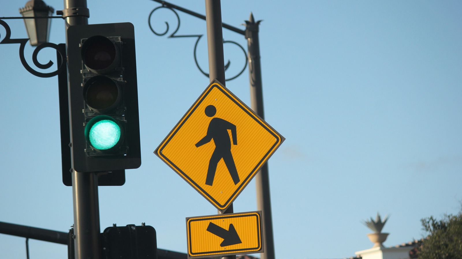 Un semáforo con la luz verde encendida junto a una señal amarilla de cruce de peatones con la figura de una persona caminando y, debajo, un letrero amarillo con una flecha, bajo un farol de hierro ornamental, con el cielo despejado y la esquina de un edificio blanco con una maceta decorativa al fondo.