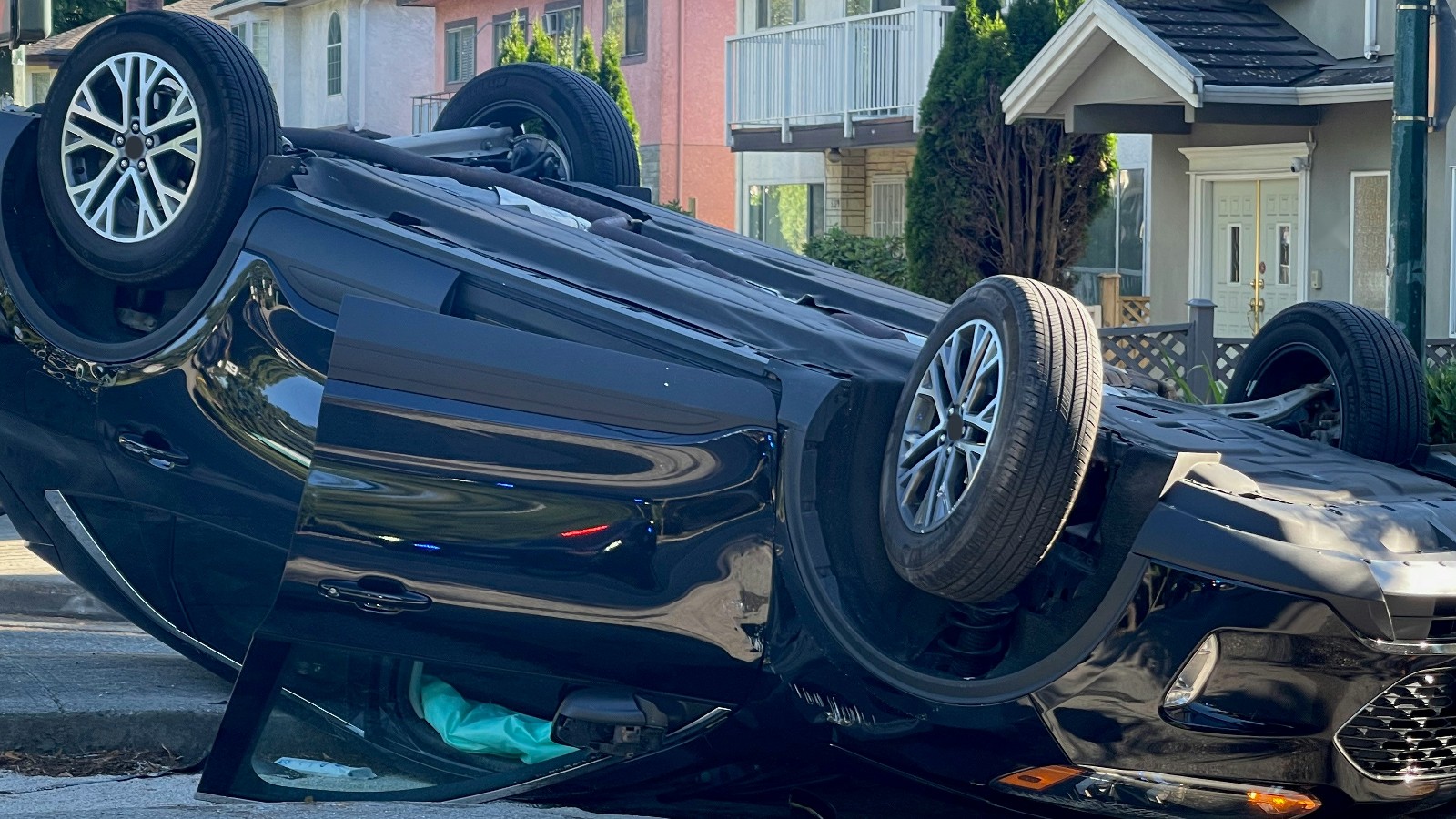 Un auto negro volcado sobre el techo junto a un bordillo, con las cuatro ruedas hacia arriba, una puerta entreabierta y una tela turquesa a la vista dentro del auto; al fondo hay casas de dos pisos, arbustos y árboles verdes.