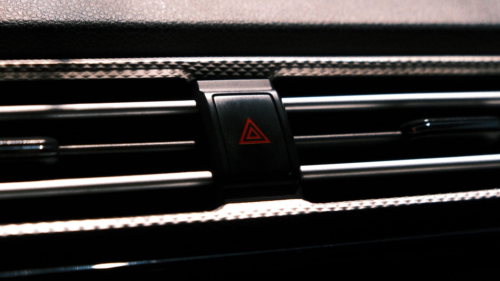 Primer plano del botón de luces de emergencia de un auto, con el triángulo rojo de advertencia, entre las rejillas de ventilación del tablero, sin personas, texto ni logotipos.