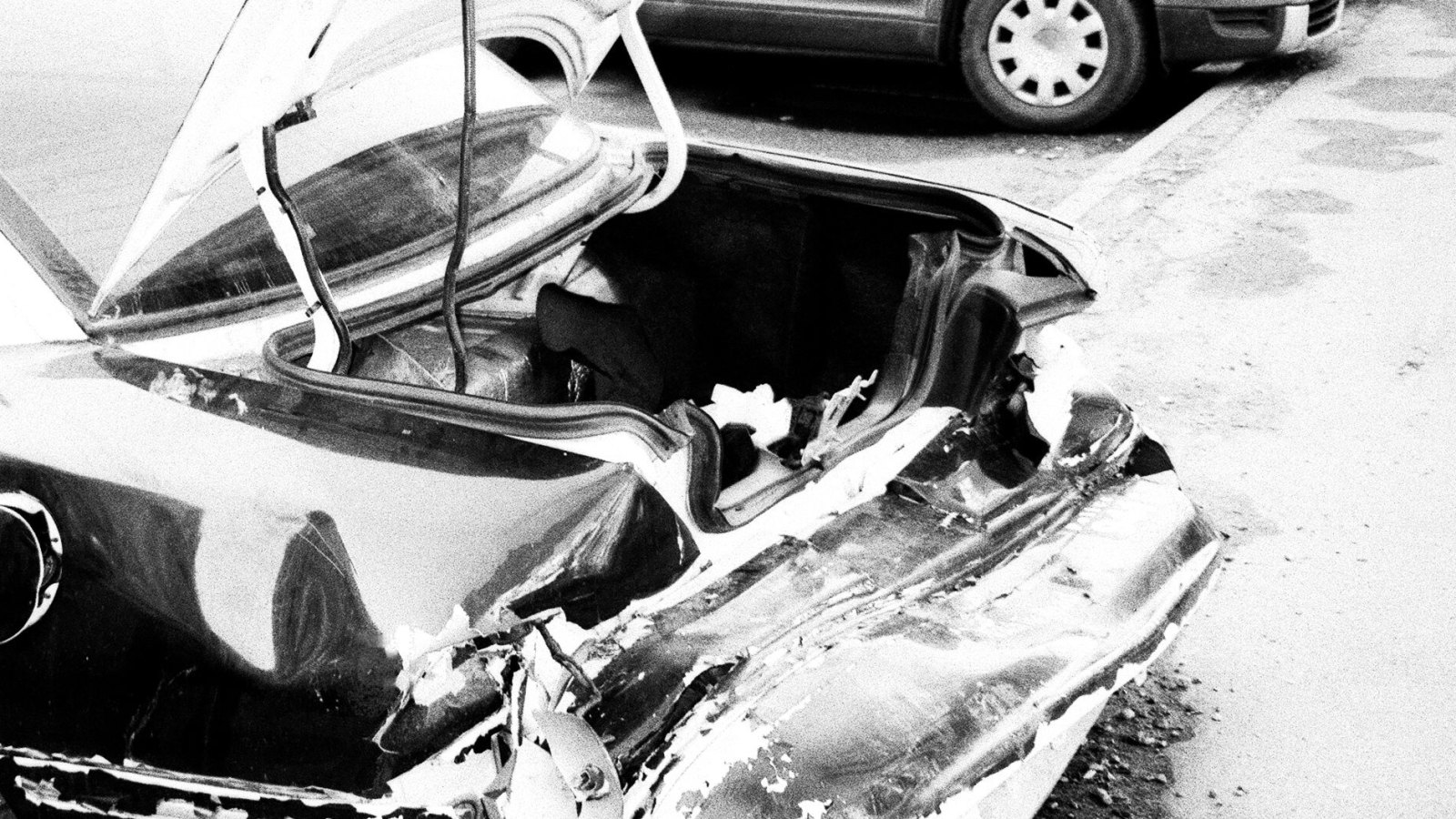 Foto en blanco y negro de la parte trasera aplastada de un sedán oscuro, con la tapa del maletero abierta y doblada, la defensa trasera arrancada y retorcida, y restos sobre el pavimento; al fondo hay otro auto estacionado junto a la banqueta.