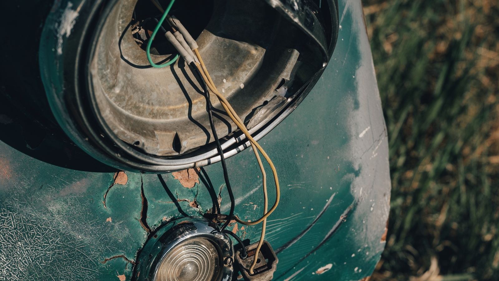 Primer plano del faro vacío y los cables expuestos de un auto dañado sobre hierba, sin personas ni señales legibles.
