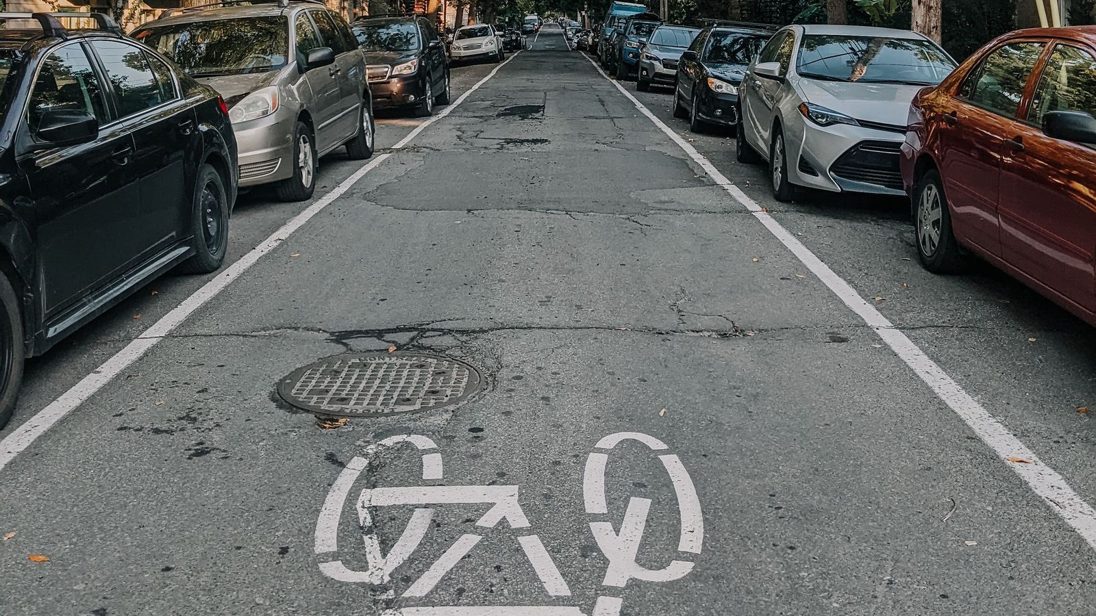 Calle con autos estacionados a ambos lados y, en el centro, un carril con el símbolo blanco de una bicicleta pintado sobre el asfalto, junto a una tapa de alcantarilla.