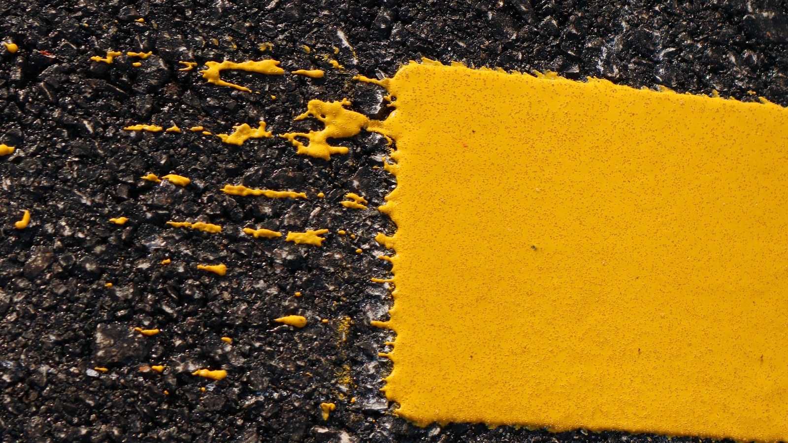 Primer plano de una franja amarilla de pintura vial sobre asfalto oscuro con salpicaduras de pintura, sin personas ni vehículos.