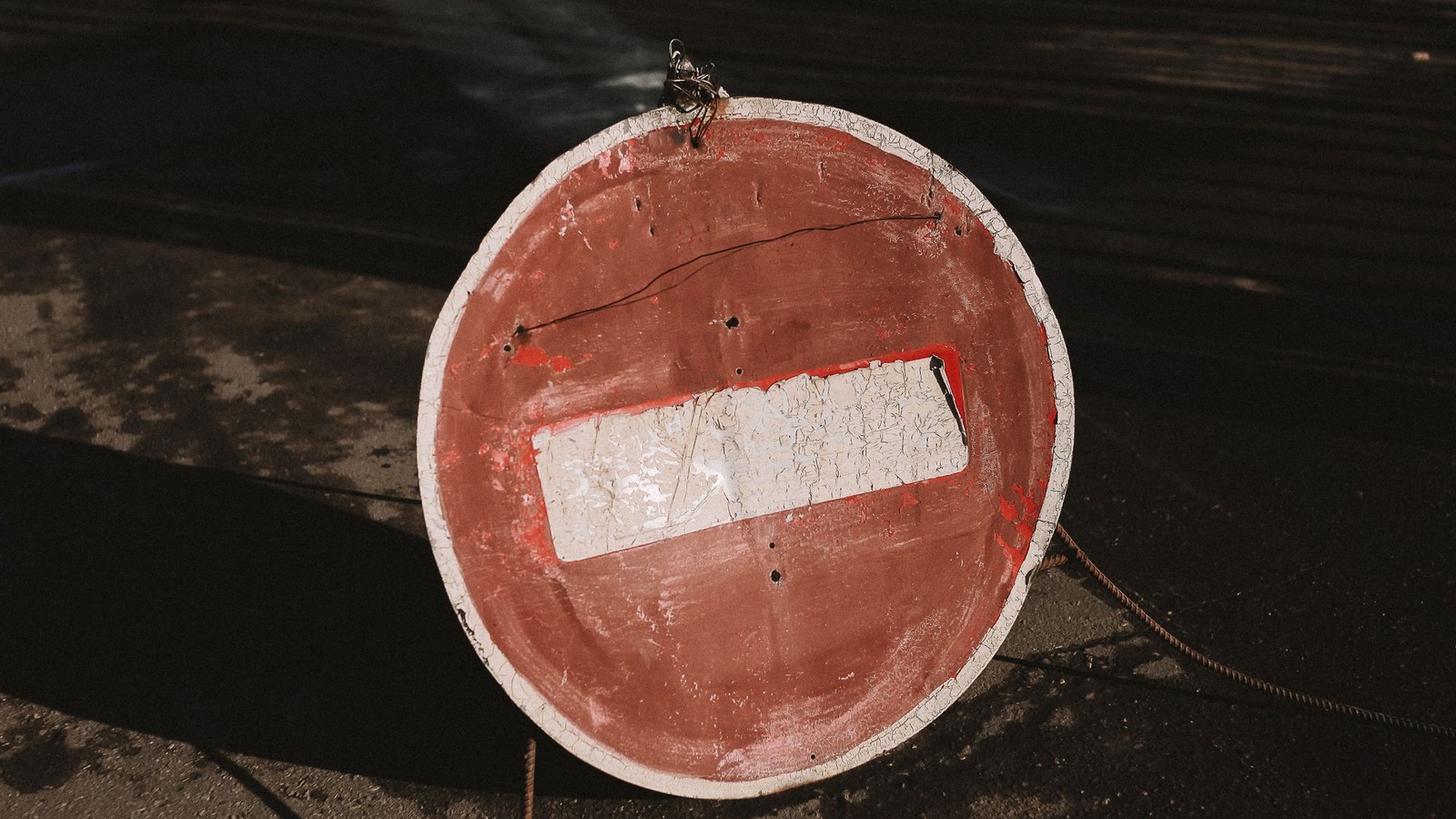 Primer plano de una señal circular de no entrar desgastada, con barra blanca sobre fondo rojo agrietado, apoyada sobre asfalto oscuro, sin personas ni texto legible.