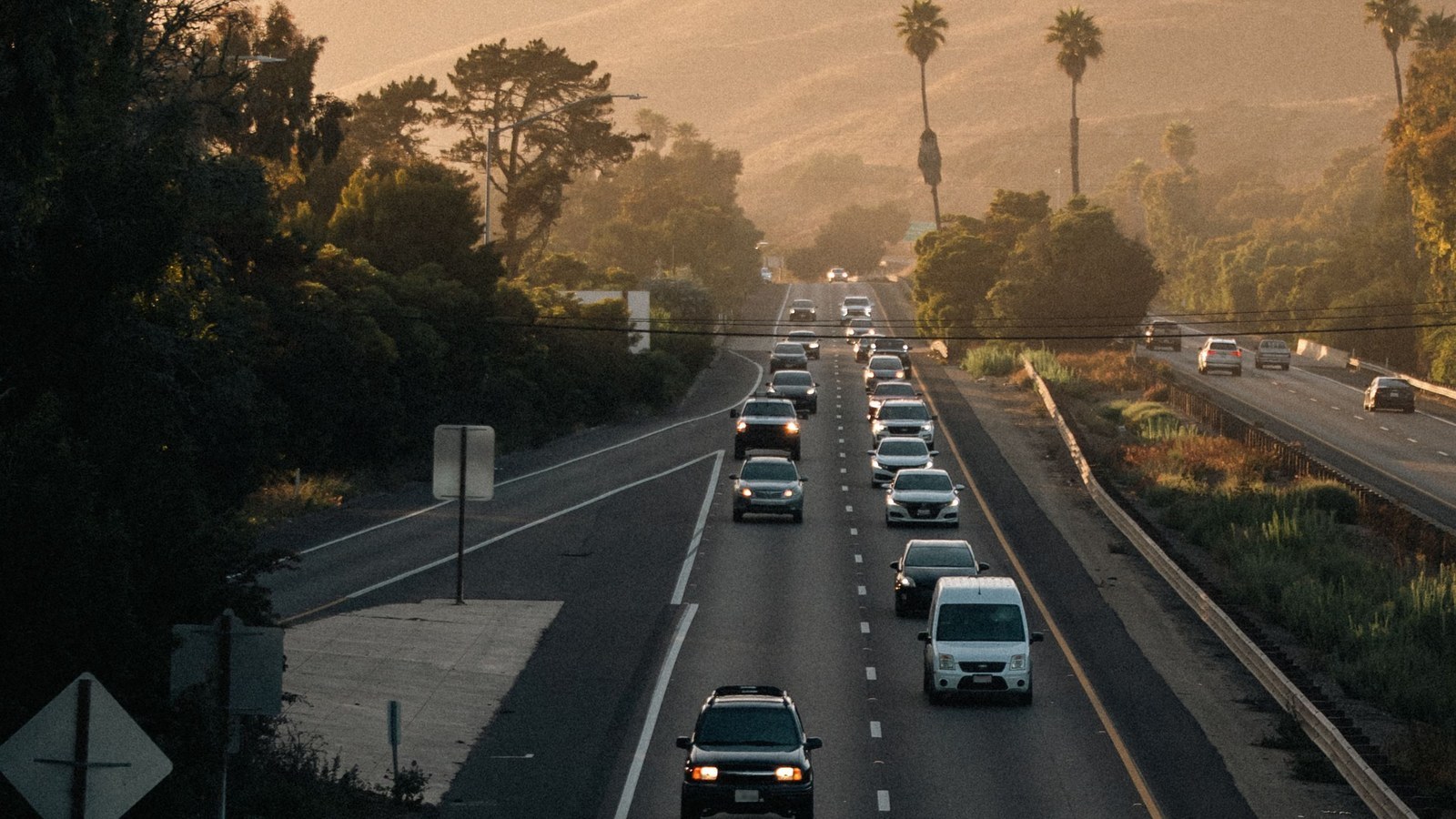 Autopista de varios carriles vista desde un paso elevado al atardecer, con autos en ambos sentidos, palmeras, árboles y colinas secas al fondo.