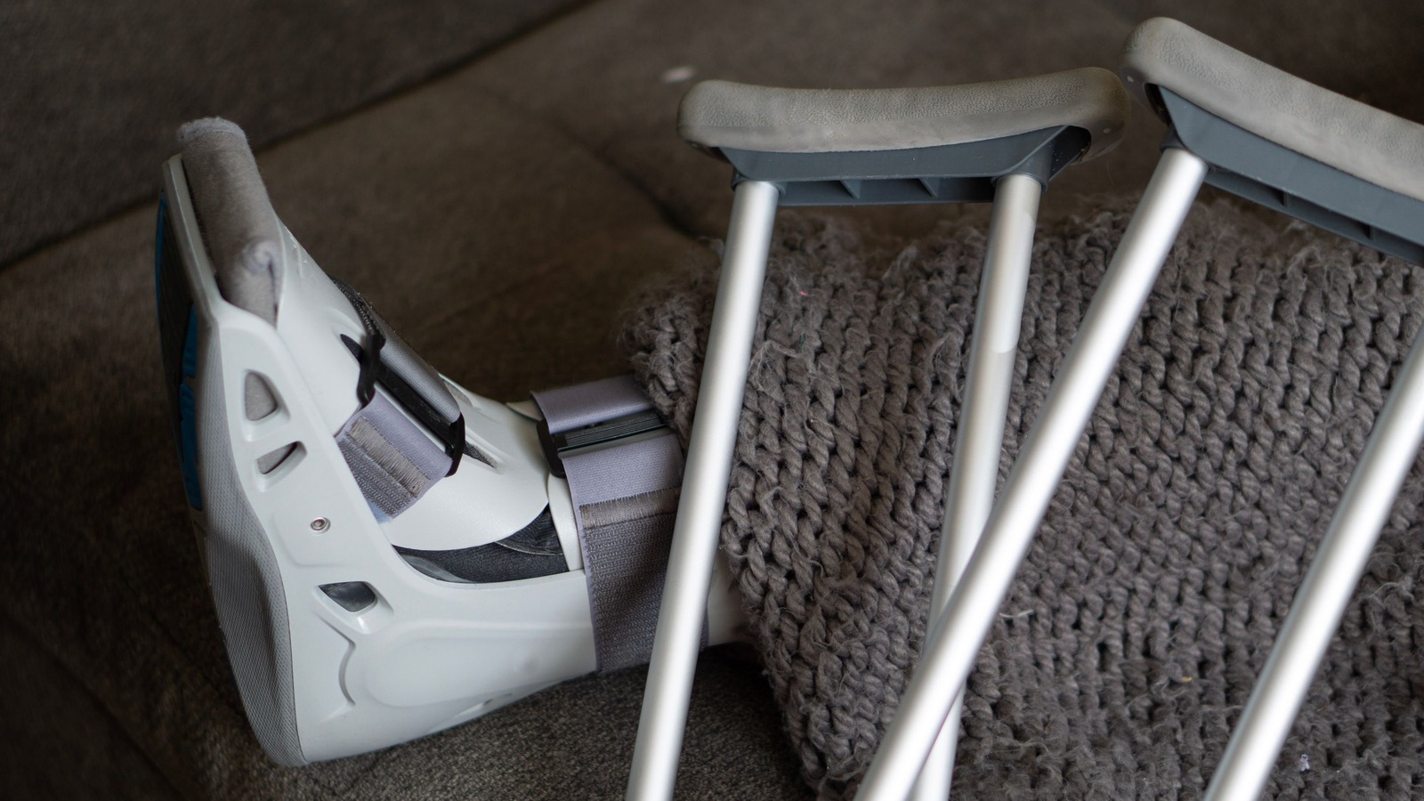 Una bota ortopédica blanca y gris con correas de velcro, apoyada sobre un sofá oscuro y cubierta en parte por una manta gris tejida, con dos muletas de aluminio recargadas encima.