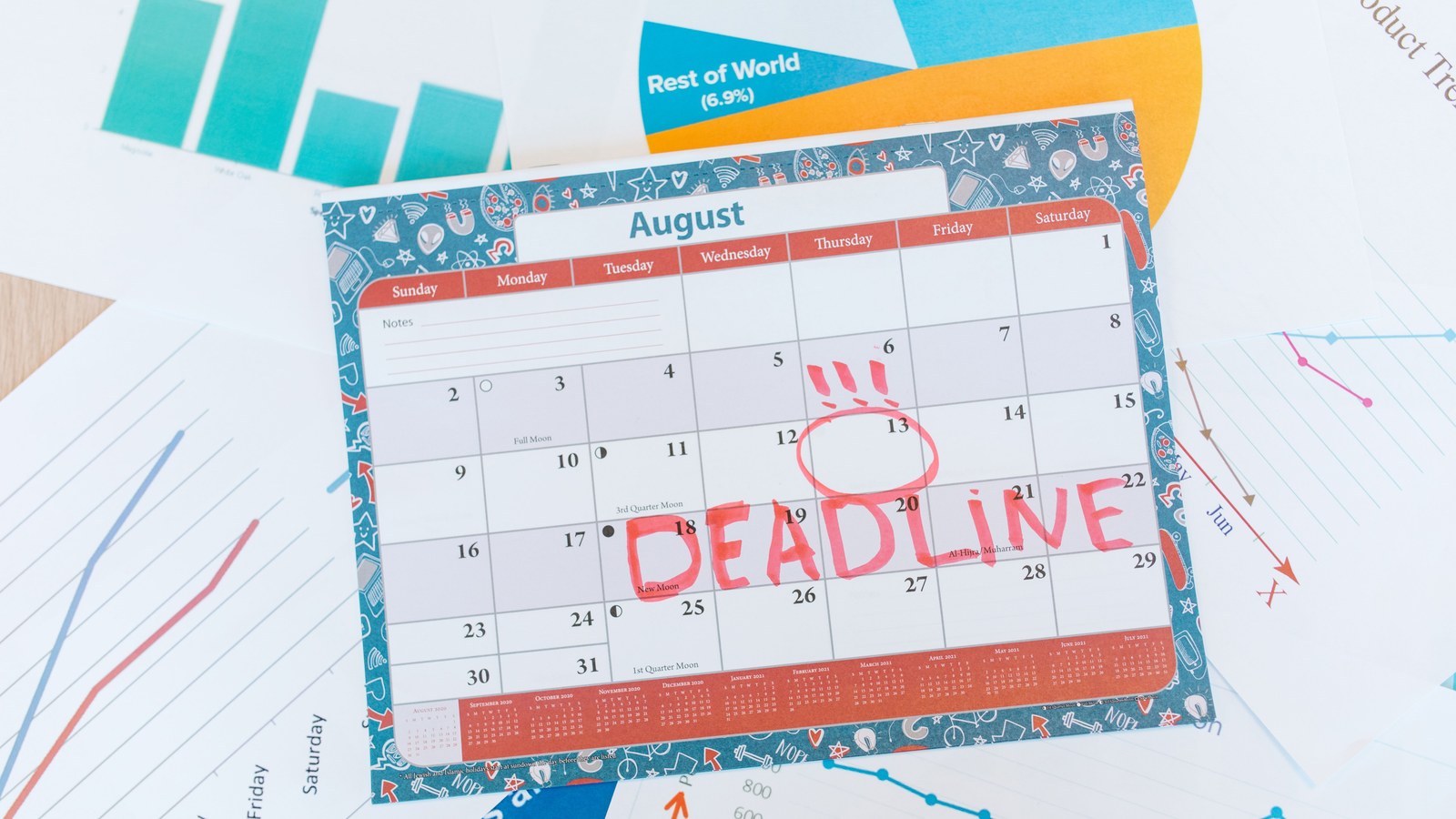 Desk calendar for August with DEADLINE written in red marker across mid-month dates and the 13th circled, resting on charts and business documents.