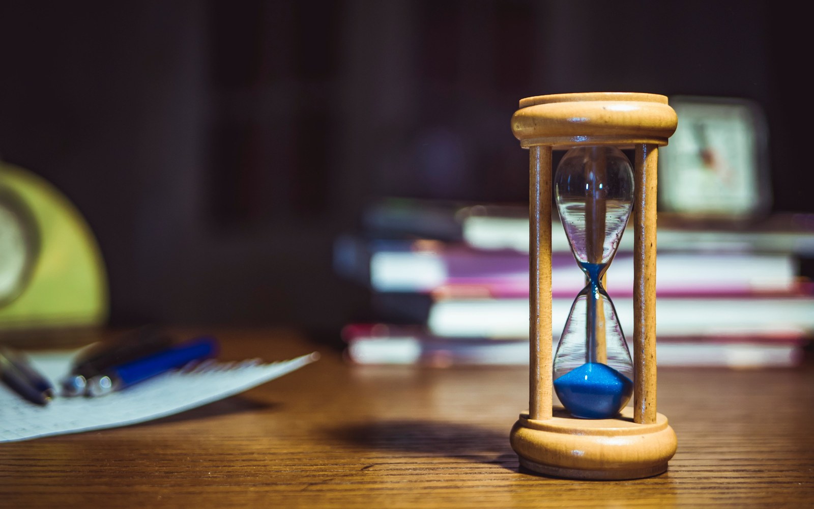 A wooden hourglass with blue sand on a desk beside a stack of books and pens, suggesting a filing deadline.