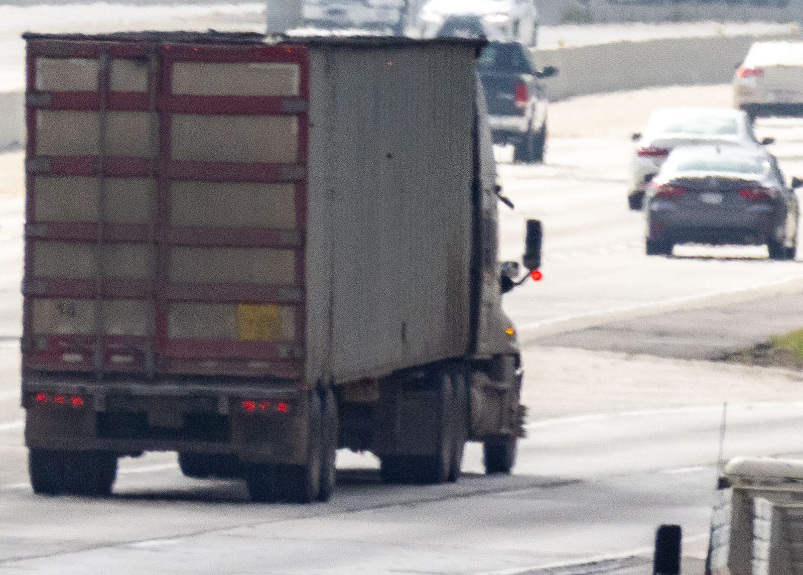 Rear view of a semi truck pulling a weathered box trailer on a busy freeway, with passenger cars in the lanes beside it.