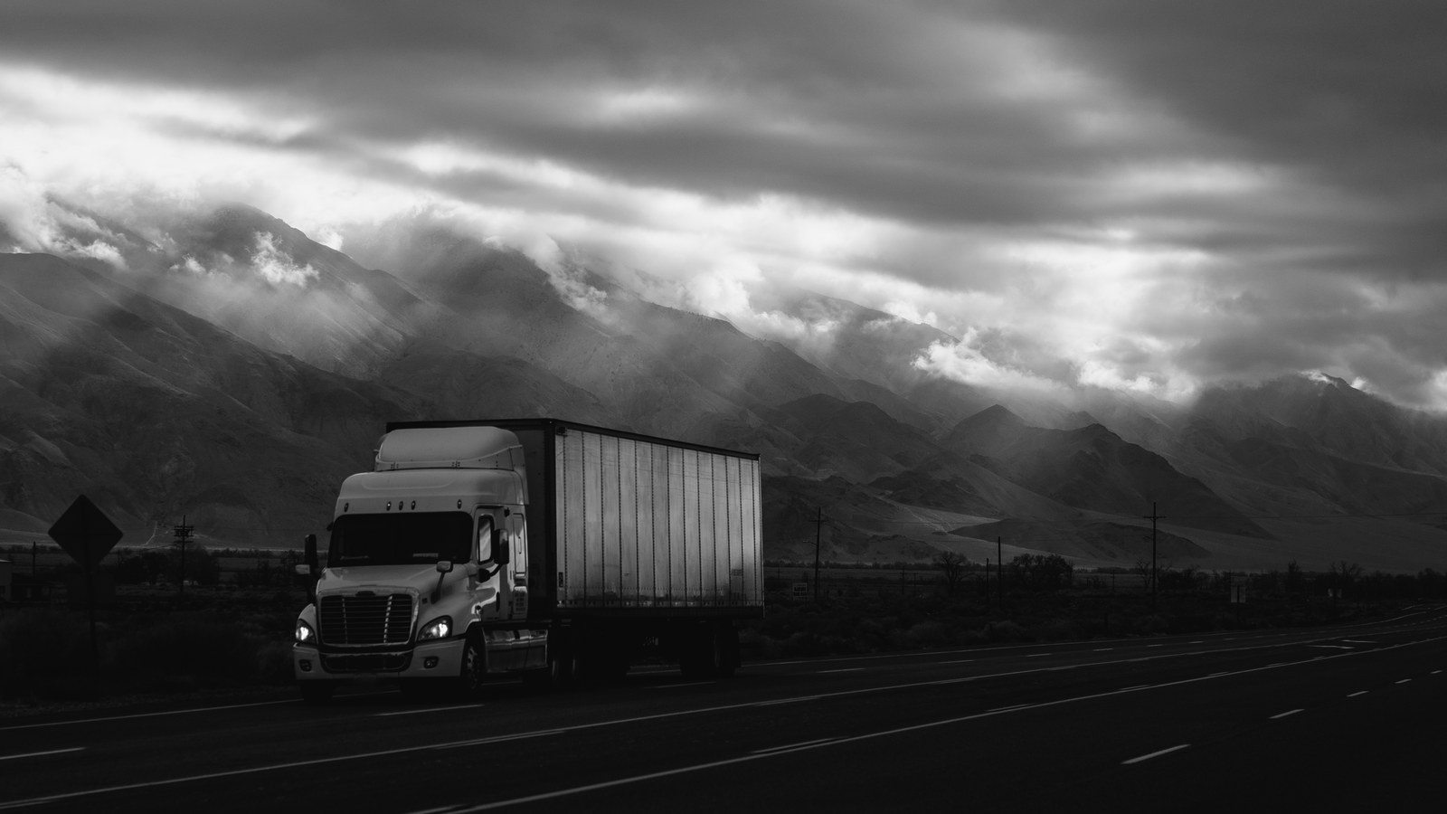 A white semi truck with its headlights on drives along a dark highway below mountains under heavy clouds, in black and white.