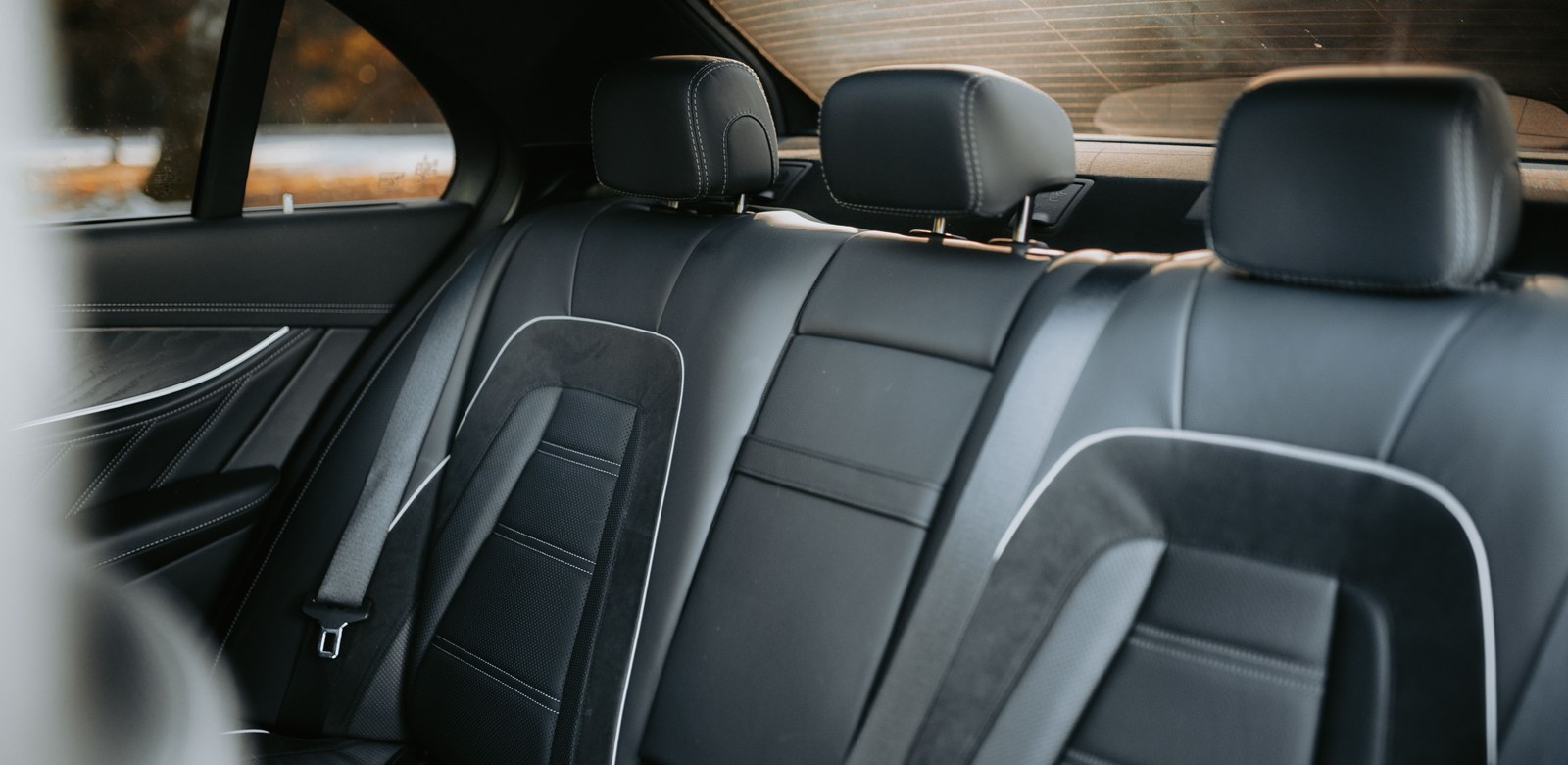 Black leather rear seat of a sedan with headrests and seat belts, seen from the back door with no passengers.